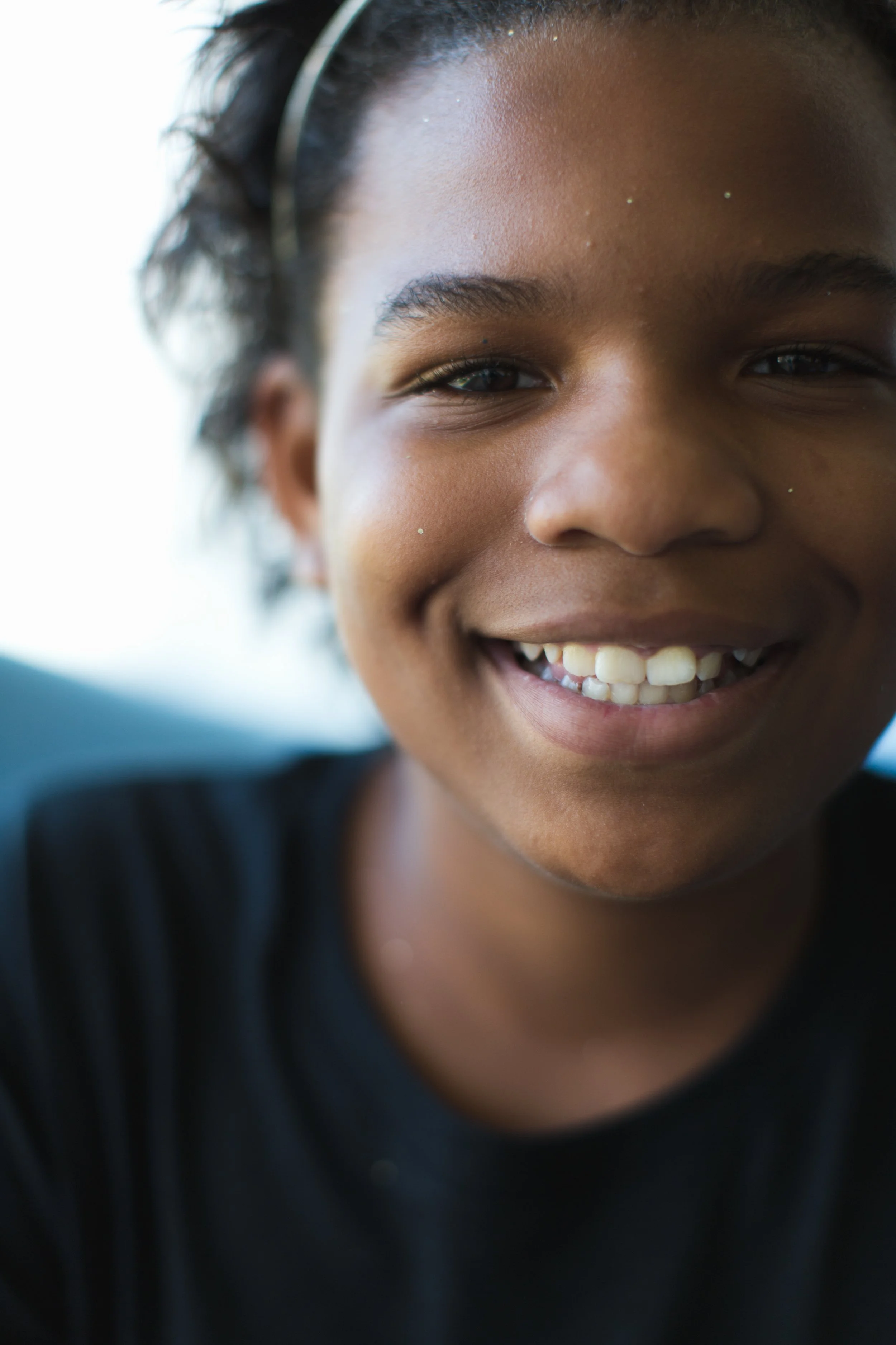 Close-up of a young person smiling, with dark hair and a bright background.