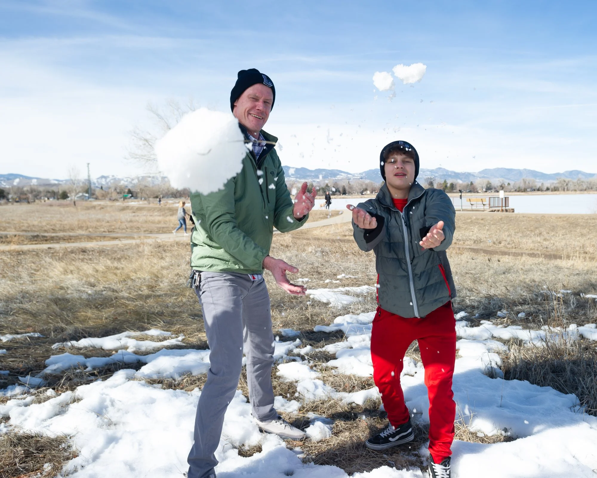 A man and a child enjoy a playful snowball fight in a snowy field. They are smiling, wearing winter clothes, with mountains in the sunny background.