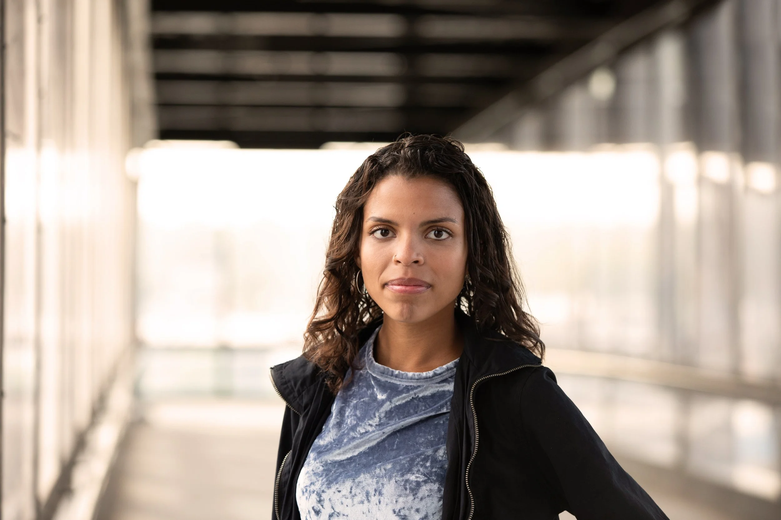 Young woman with dark curly hair, wearing a jacket and a patterned shirt, standing in an urban subway or train station with a blurred tunnel background.