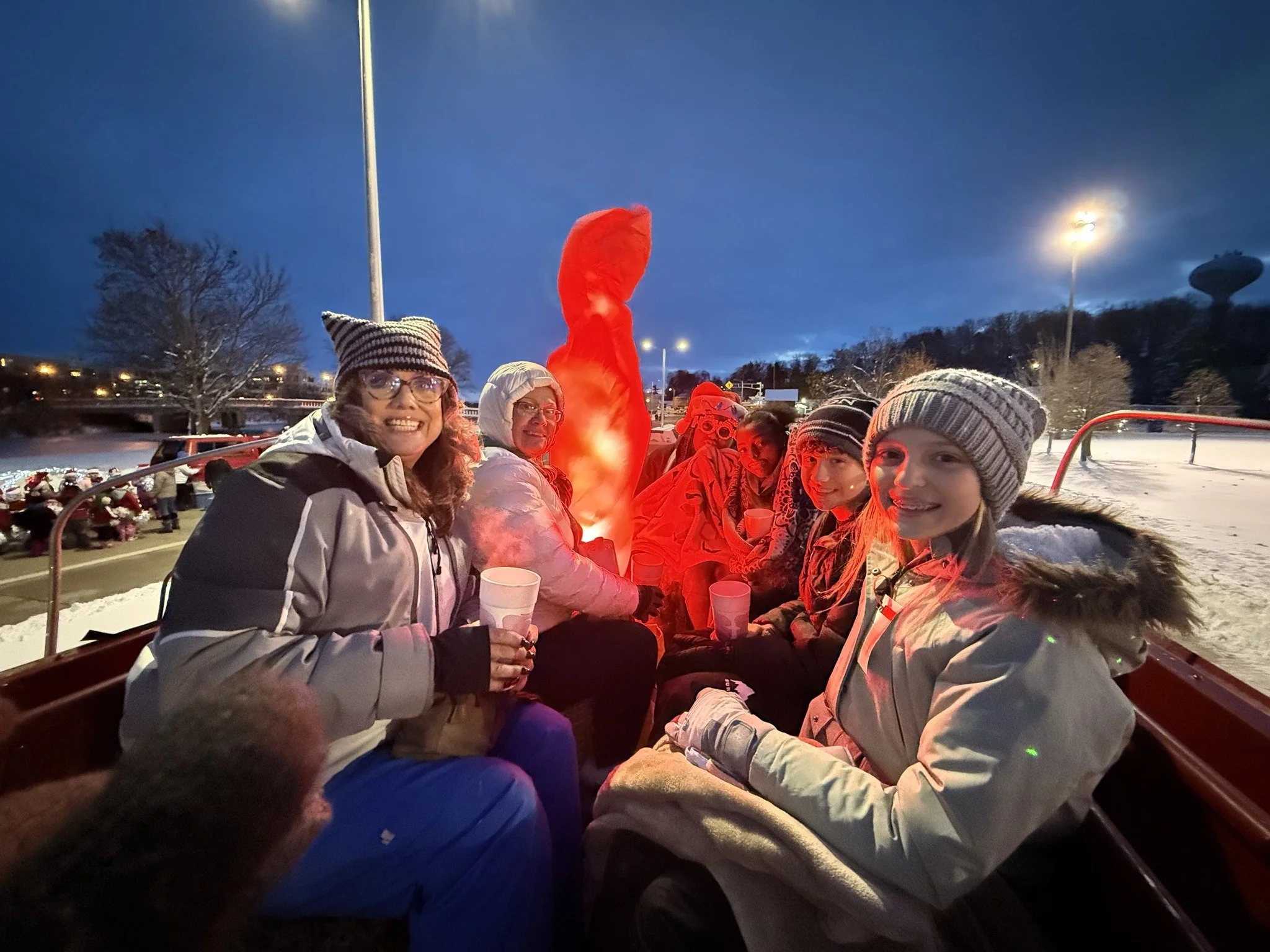 A group of smiling people bundled in winter clothing, holding cups, sits on a festive open-air ride under streetlights and a dusky sky, conveying warmth and camaraderie.