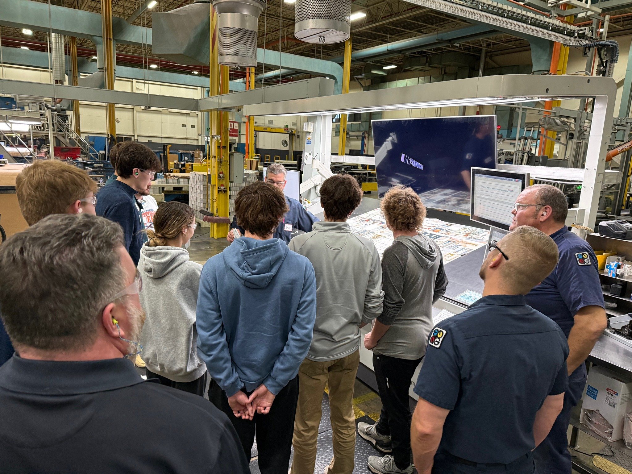 A group of people gather around a printing press in a factory, attentively watching a demonstration. The setting is industrial with machinery and monitors.