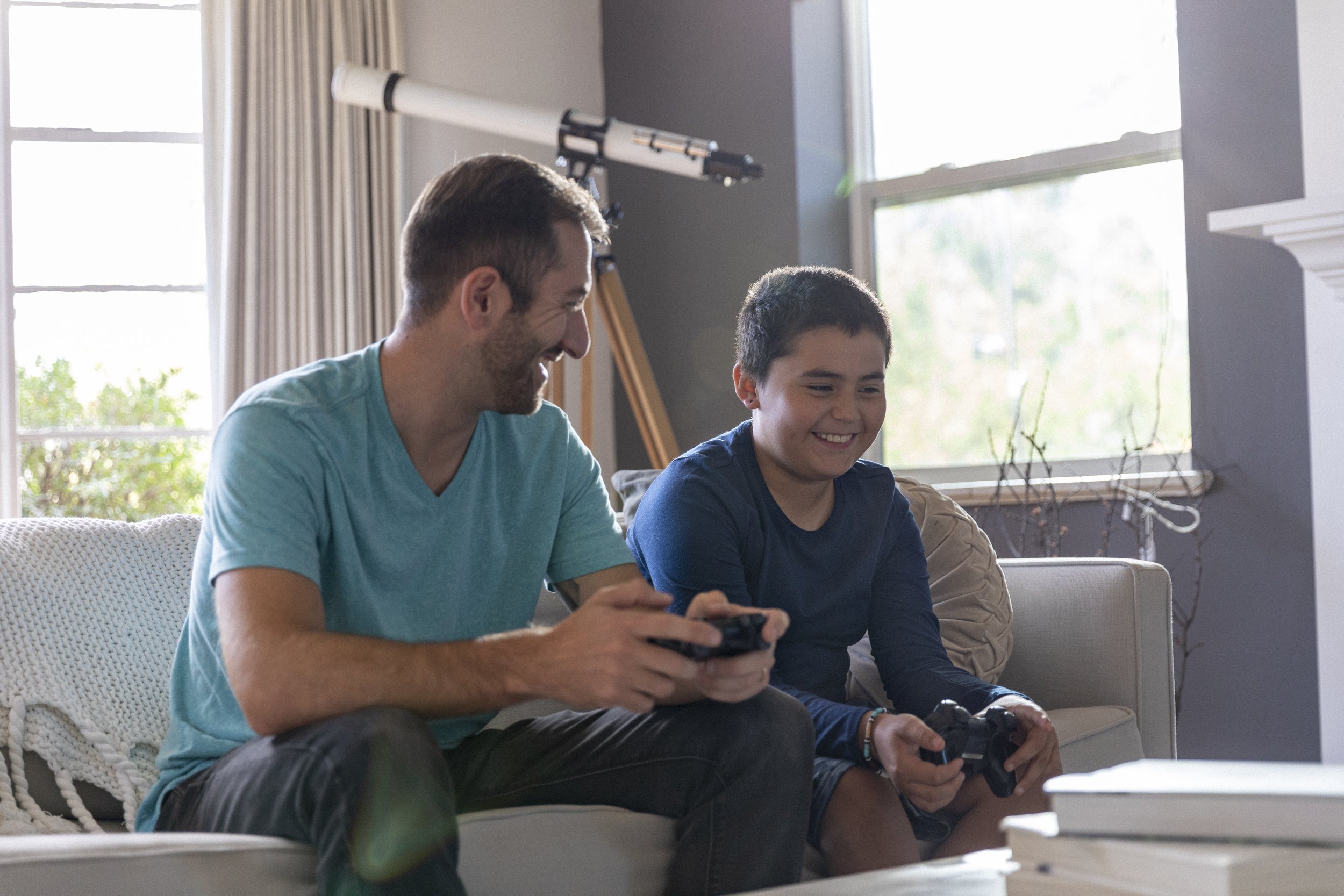 A man and a boy sitting on a beige couch in a bright living room, playing video games together, smiling.