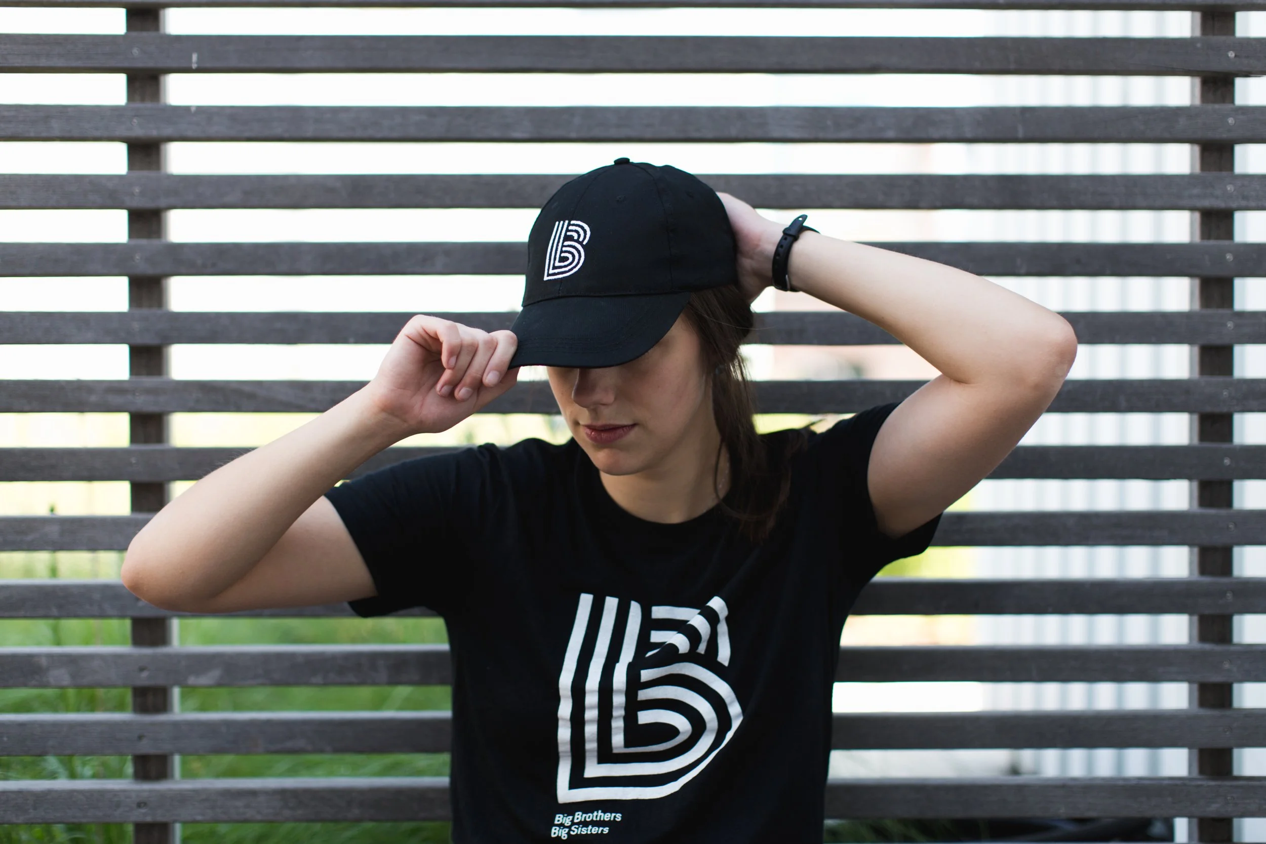 A woman wearing a black cap and black t-shirt with a big white logo, adjusting her cap with her right hand, standing in front of a wooden slat fence.