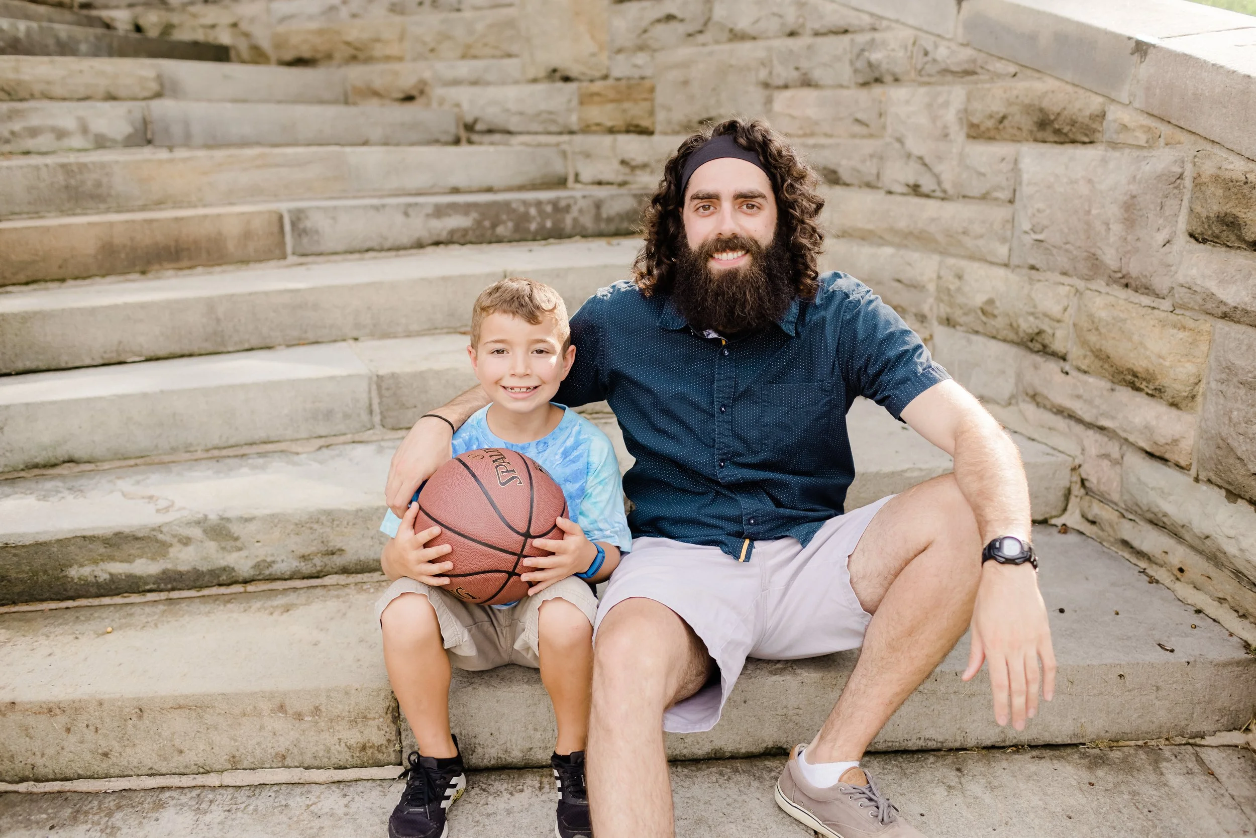 A man with long curly hair and a beard sitting on outdoor stone steps next to a young boy holding a basketball. The man is smiling and wearing a dark blue shirt, light shorts, and a watch. The boy, wearing a blue T-shirt, khaki shorts, and black shoes, is also smiling.