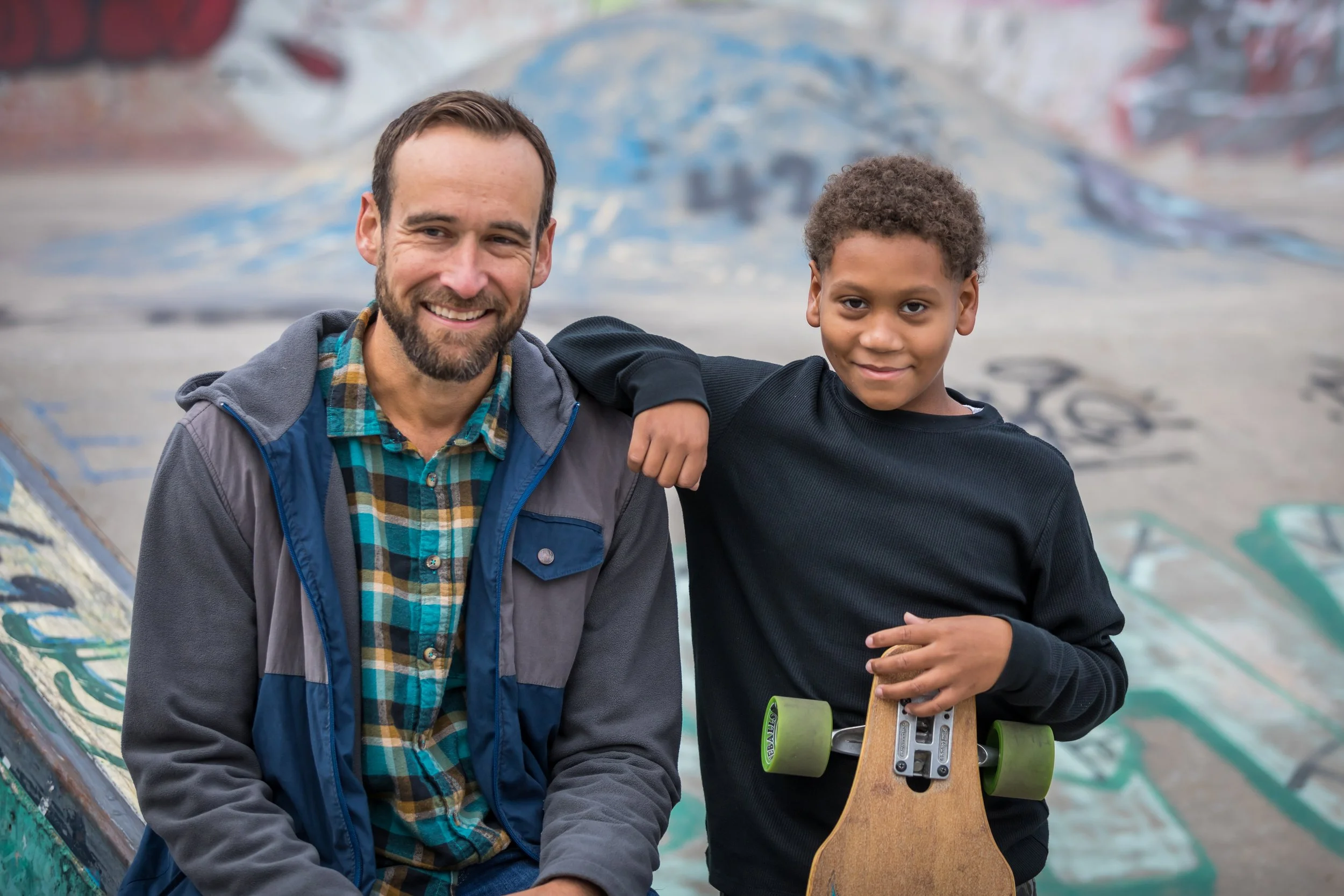 A smiling young boy poses at a skate park. The boy holds a skateboard. Graffiti decorates the ramps behind him. He appears happy and relaxed.