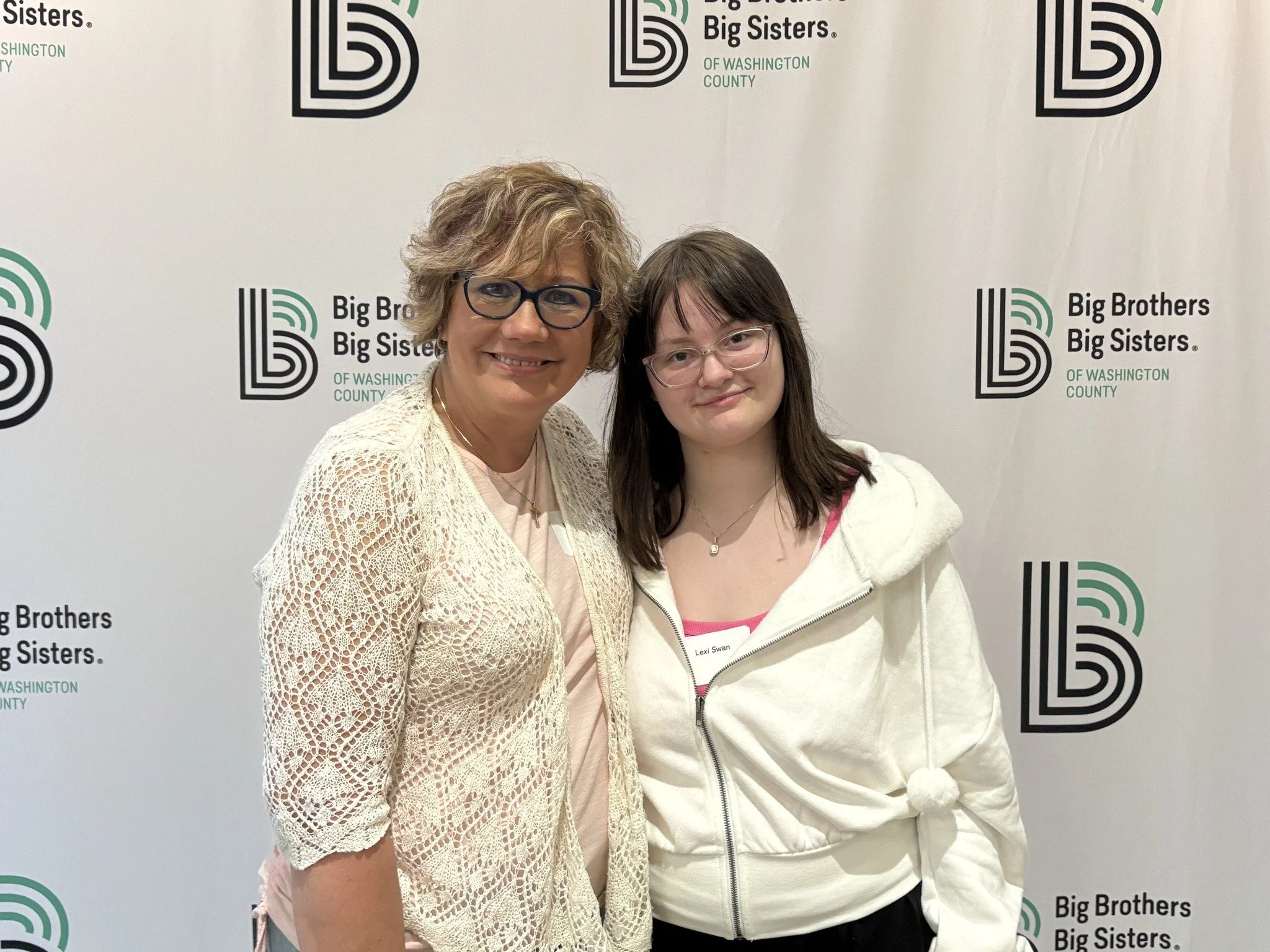 Two women posing together in front of a backdrop for Big Brothers Big Sisters of Washington County. The woman on the left has curly, light brown hair, glasses, and is wearing a cream-colored lace cardigan over a pink top. The woman on the right has straight, dark brown hair, glasses, and is wearing a white hoodie with a pink trim.