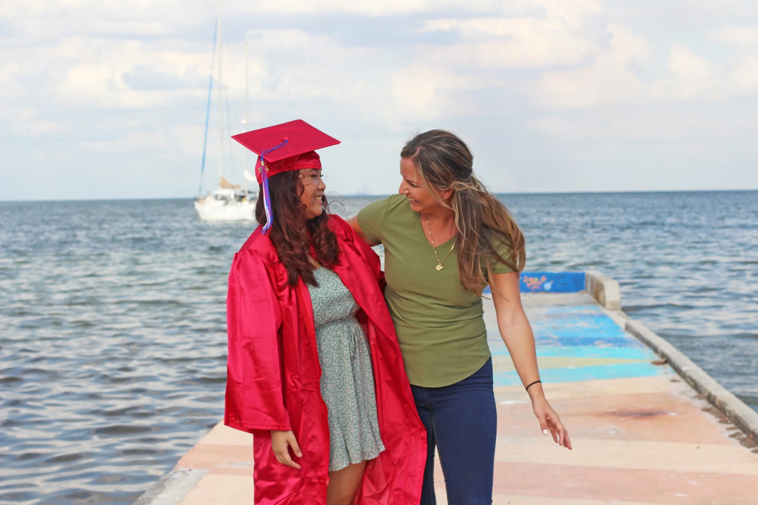 Two women walk arm in arm on a pier by the sea. One is wearing a red graduation gown and cap, while the other is in casual clothes, smiling warmly.