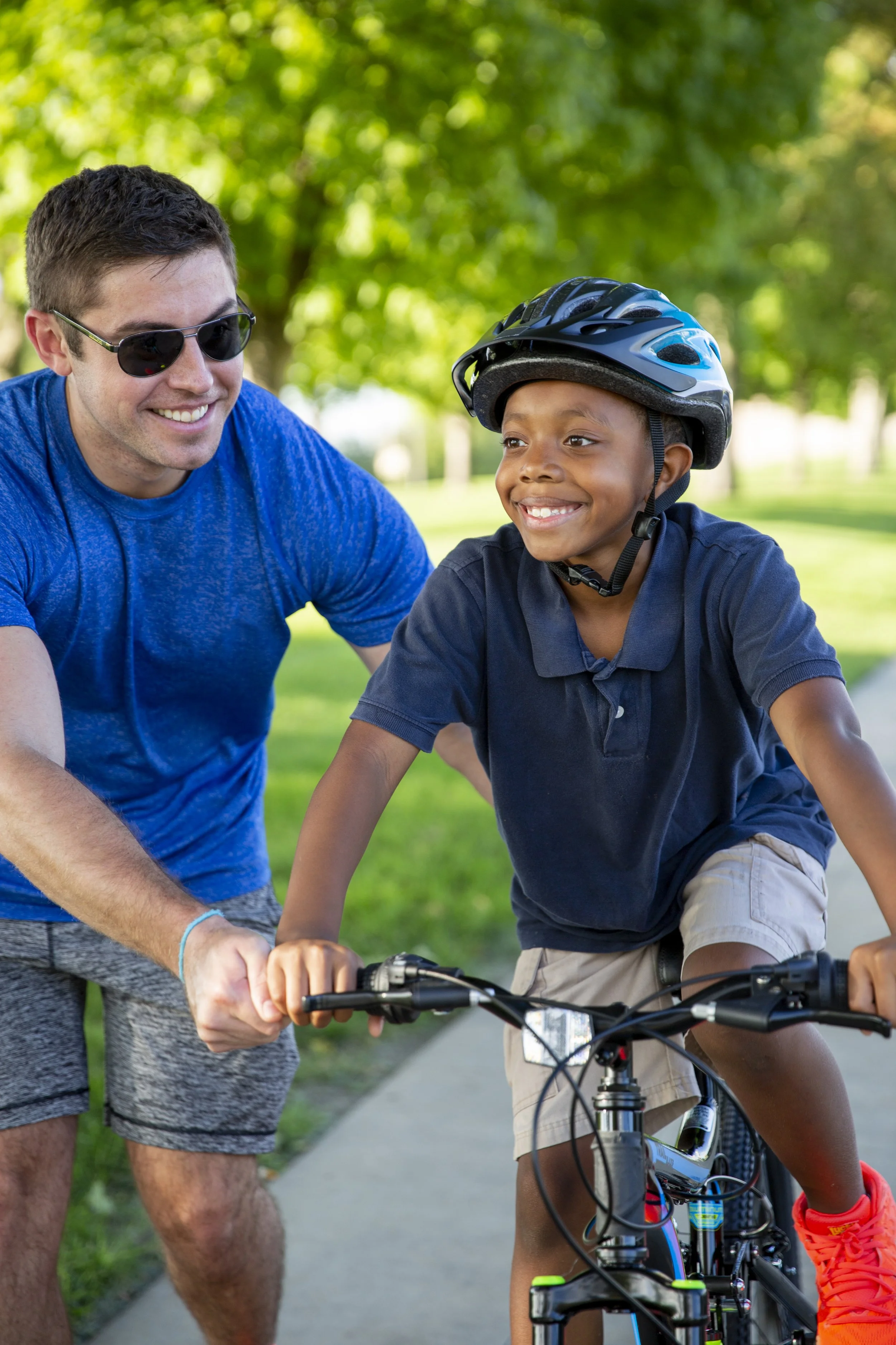 A man in sunglasses assists a smiling boy riding a bicycle on a sunny day in a park. The boy wears a helmet, conveying a sense of joy and learning.