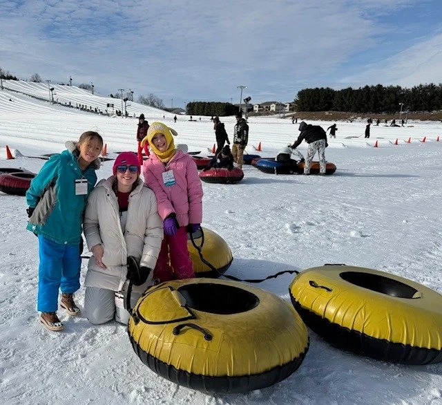 Three people, dressed in colorful winter clothing, smile near yellow snow tubes on a snowy slope. Others in the background are preparing to snow tube.