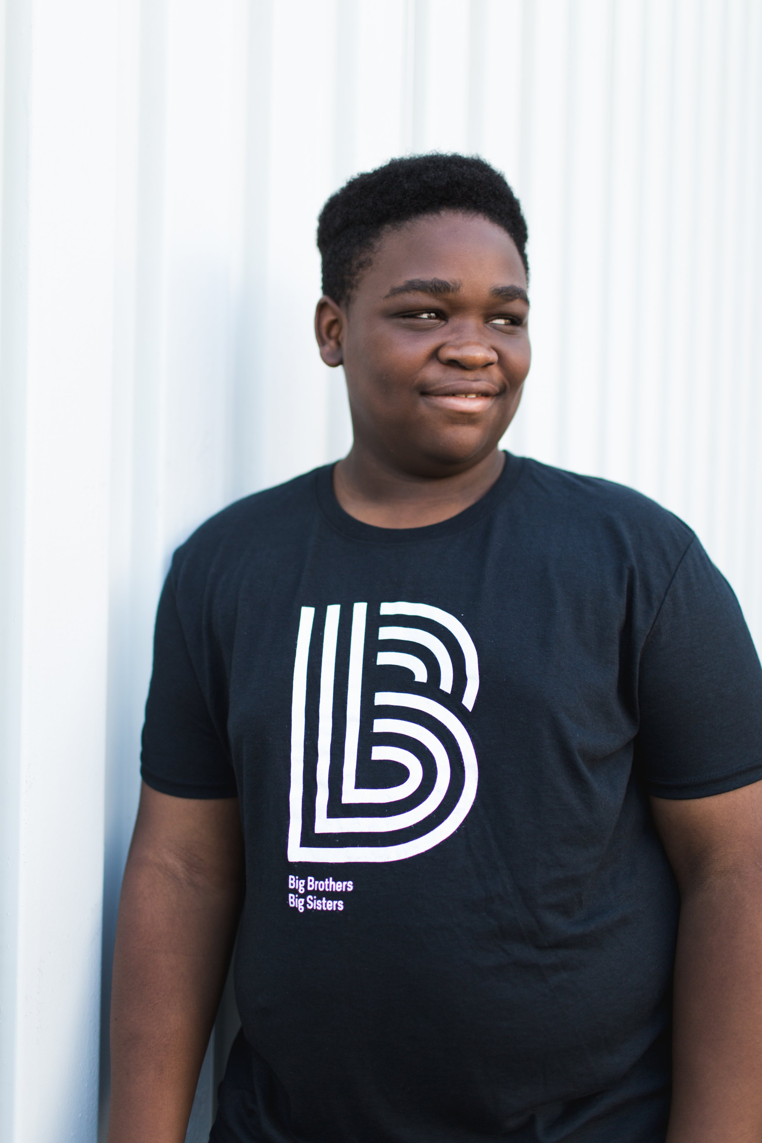 A young African American person with short curly hair, wearing a black T-shirt with a white line art design and the words "Big Brothers Big Sisters," standing against a light-colored, vertically-striped background, looking to the side and smiling subtly.