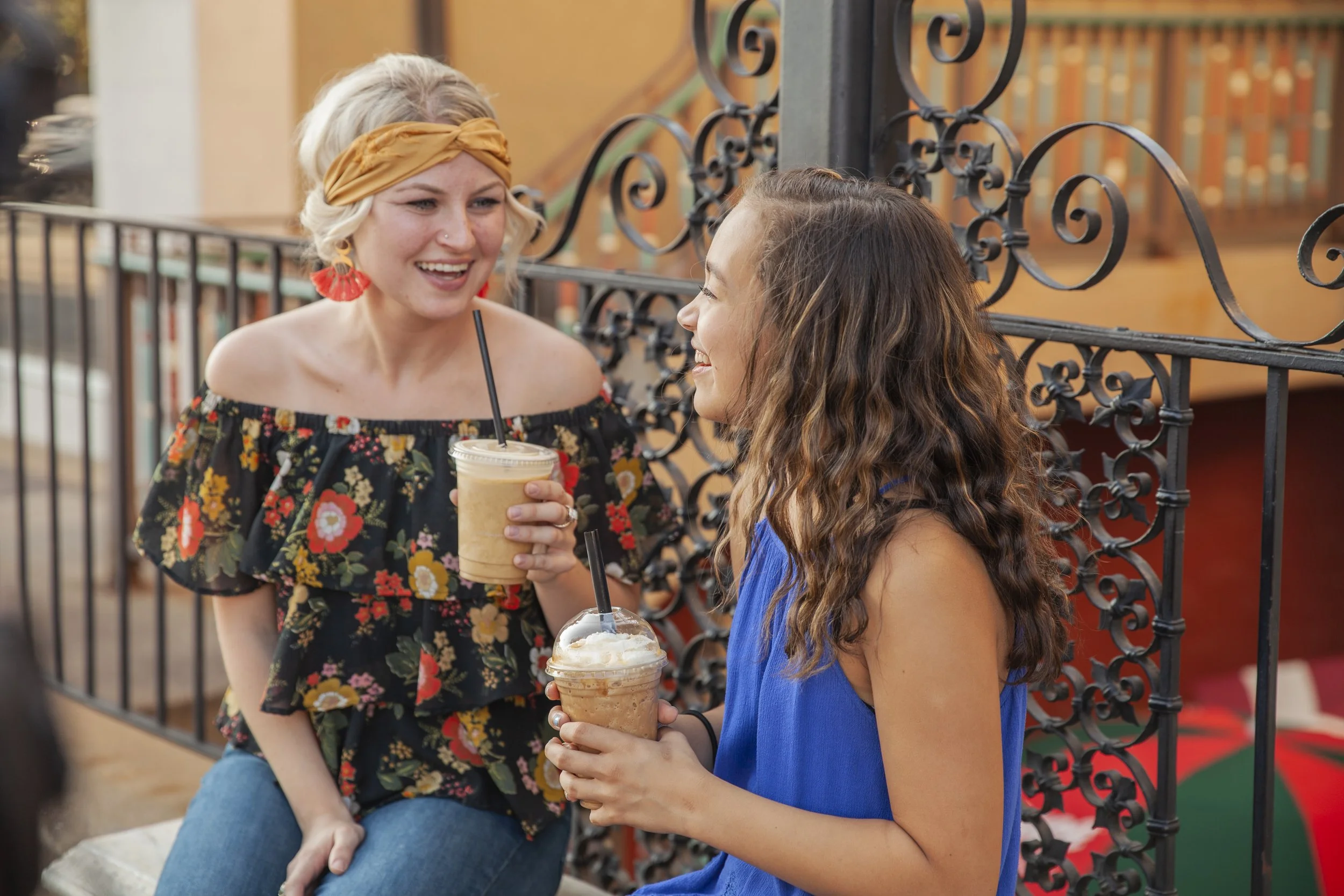 Two women sit outdoors, happily chatting over iced coffees. One wears a floral top and yellow headband, the other a blue dress. The scene is warm and cheerful.