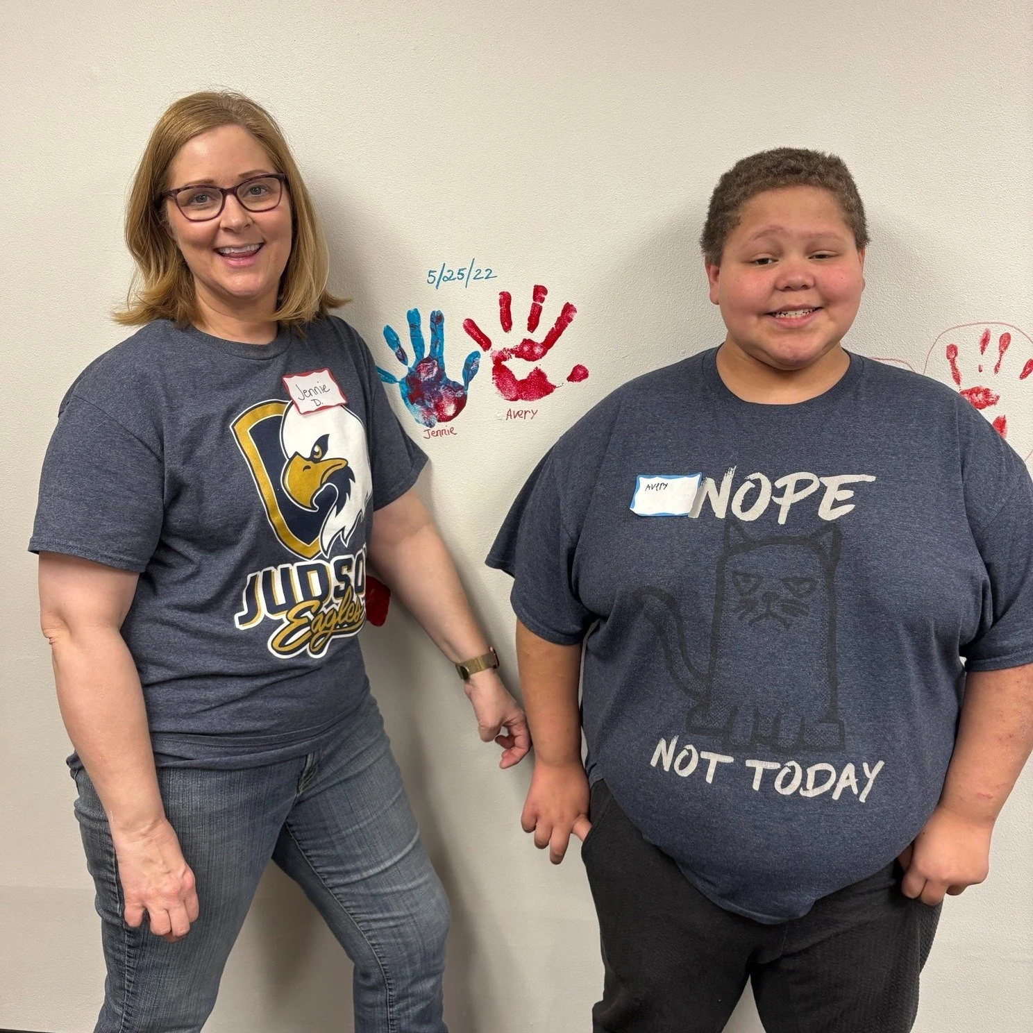 Two people smiling in front of a wall with colorful handprints and dates.