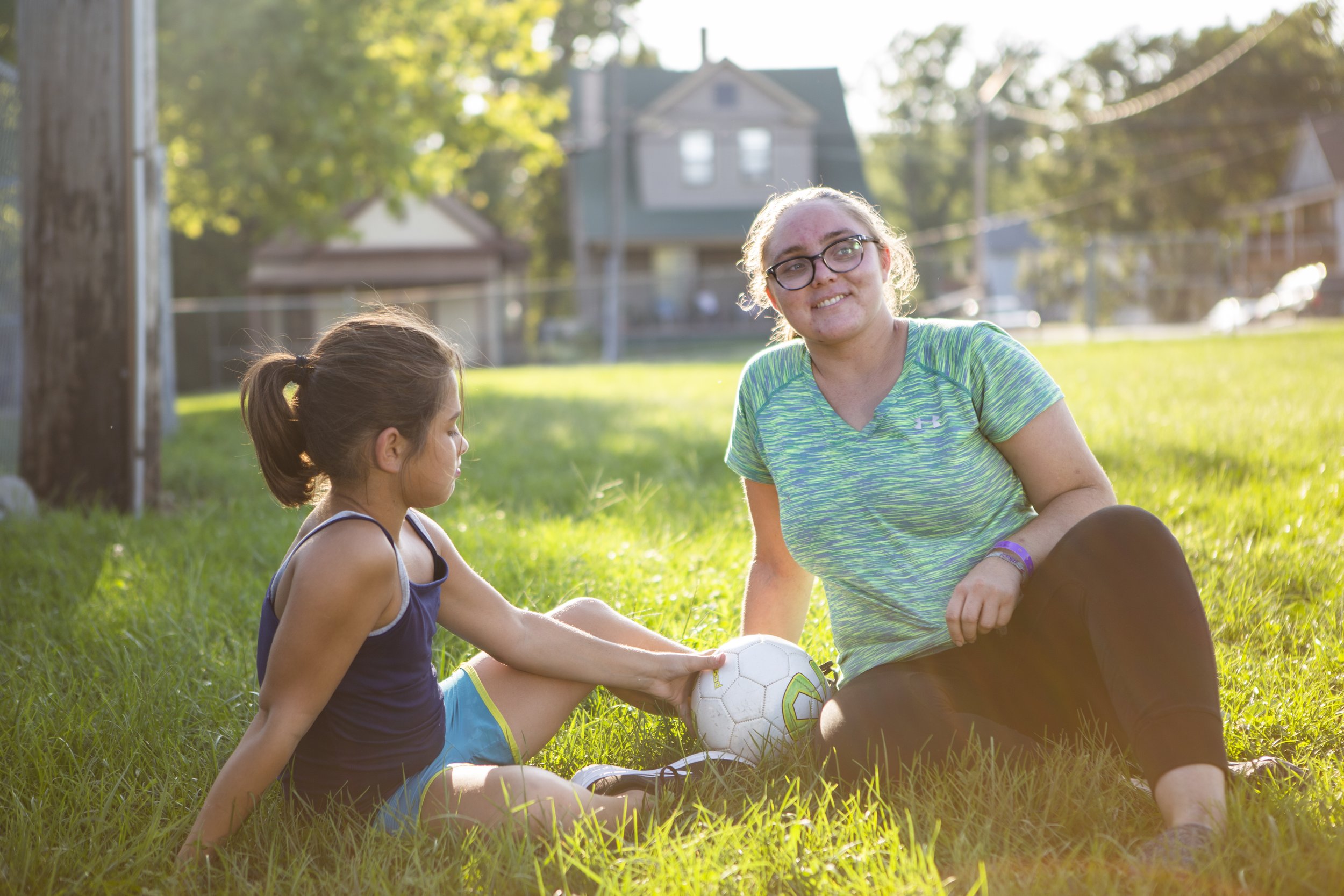A young girl and an adult woman sitting on grass in a park, with a soccer ball between them, during daytime. The girl is in a blue tank top, and the woman is wearing glasses and a green striped shirt. They are smiling and enjoying the sunny outdoor environment with trees and houses in the background.