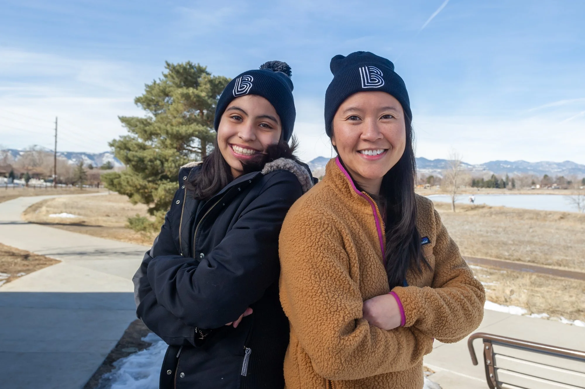 Two women smiling with arms crossed, wearing matching winter hats and coats, stand outdoors with a scenic snowy landscape and mountains in the background.