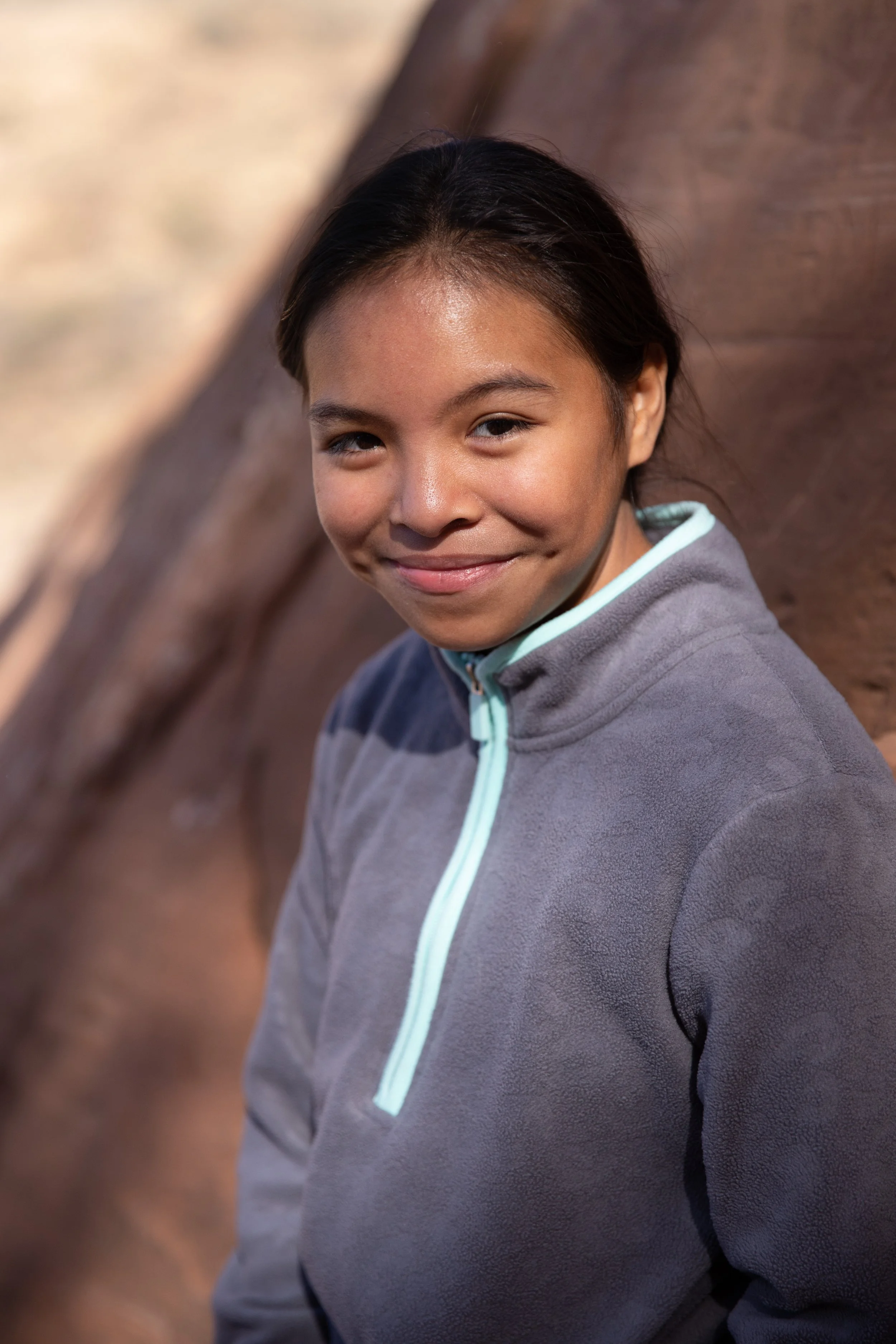 A young girl with long dark hair, smiling with a closed-mouth smile, wearing a gray zip-up jacket with a light blue zipper, standing outdoors near a rocky background.