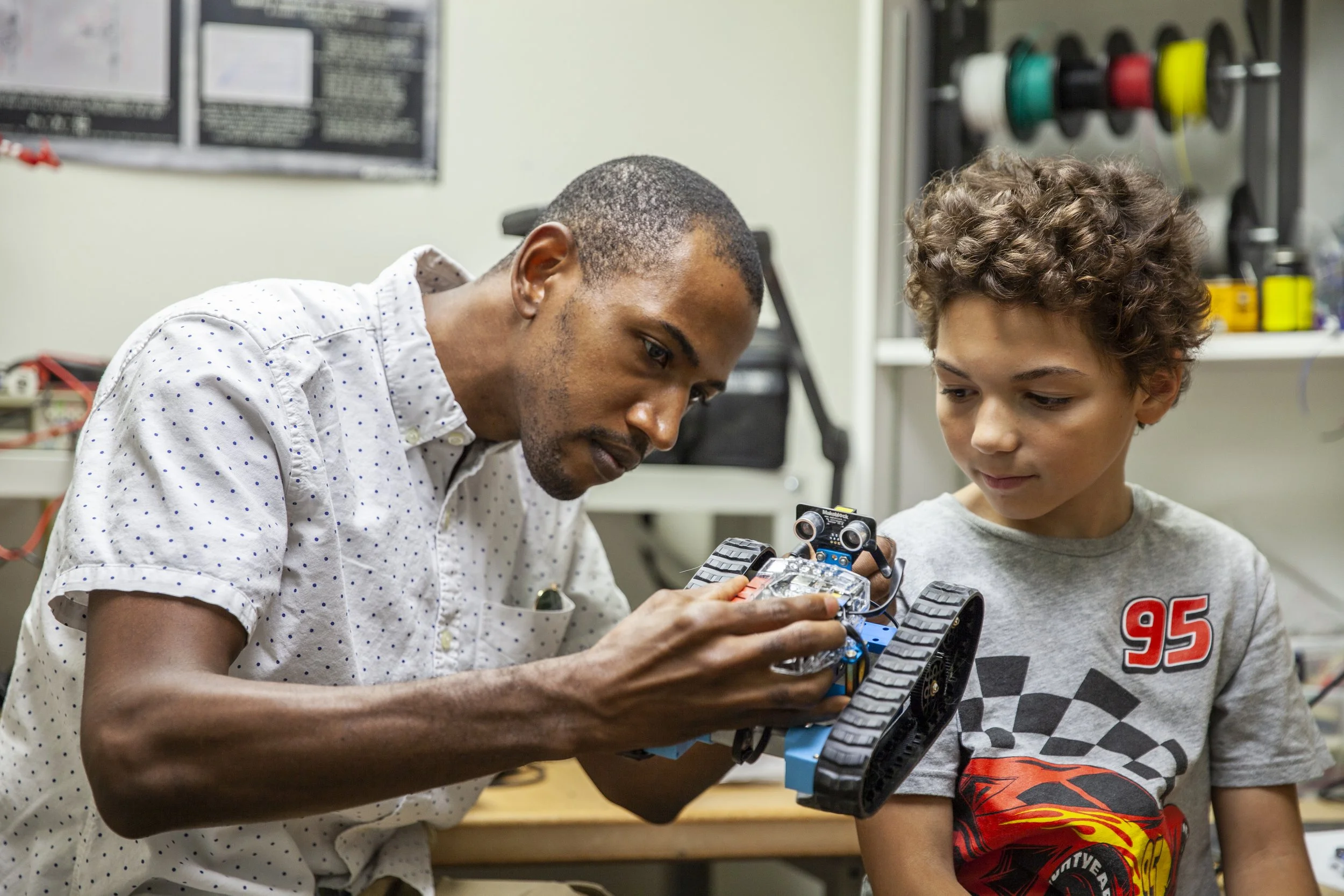 A man in a white shirt shows a young boy in a racing-themed shirt how to assemble a robotic car in a workshop. They focus intently, conveying collaboration.