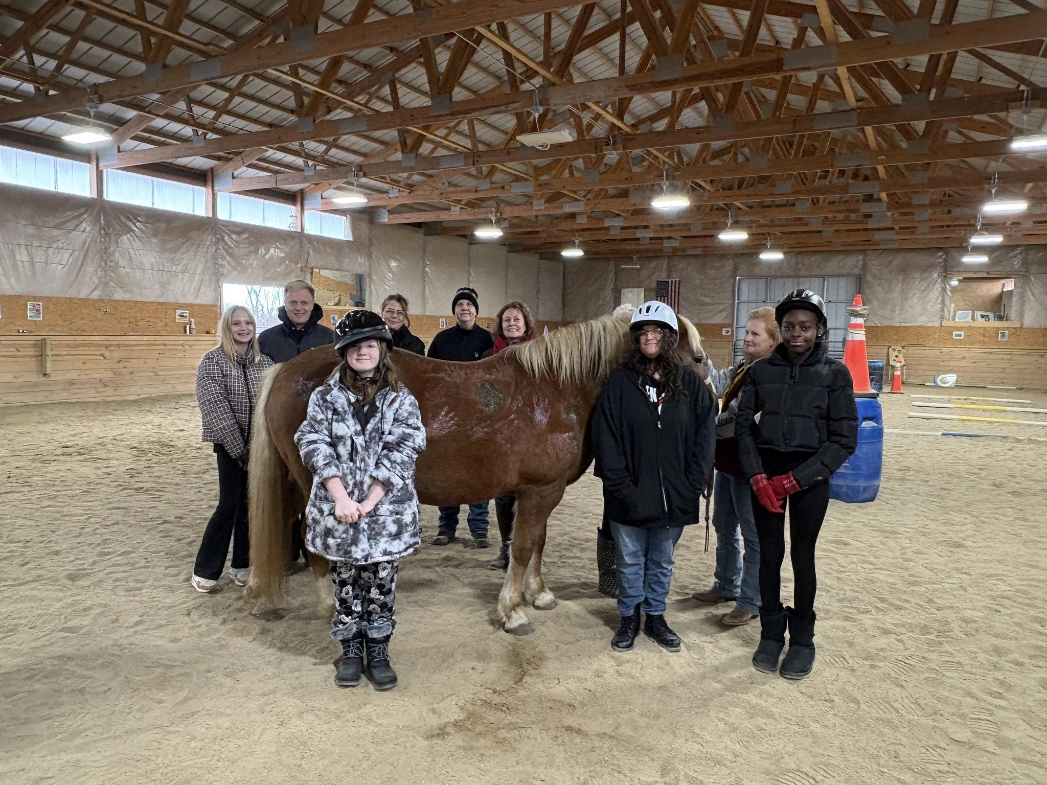 A group of people, including children wearing helmets, stand in an indoor arena with a brown horse. The mood is cheerful and supportive.