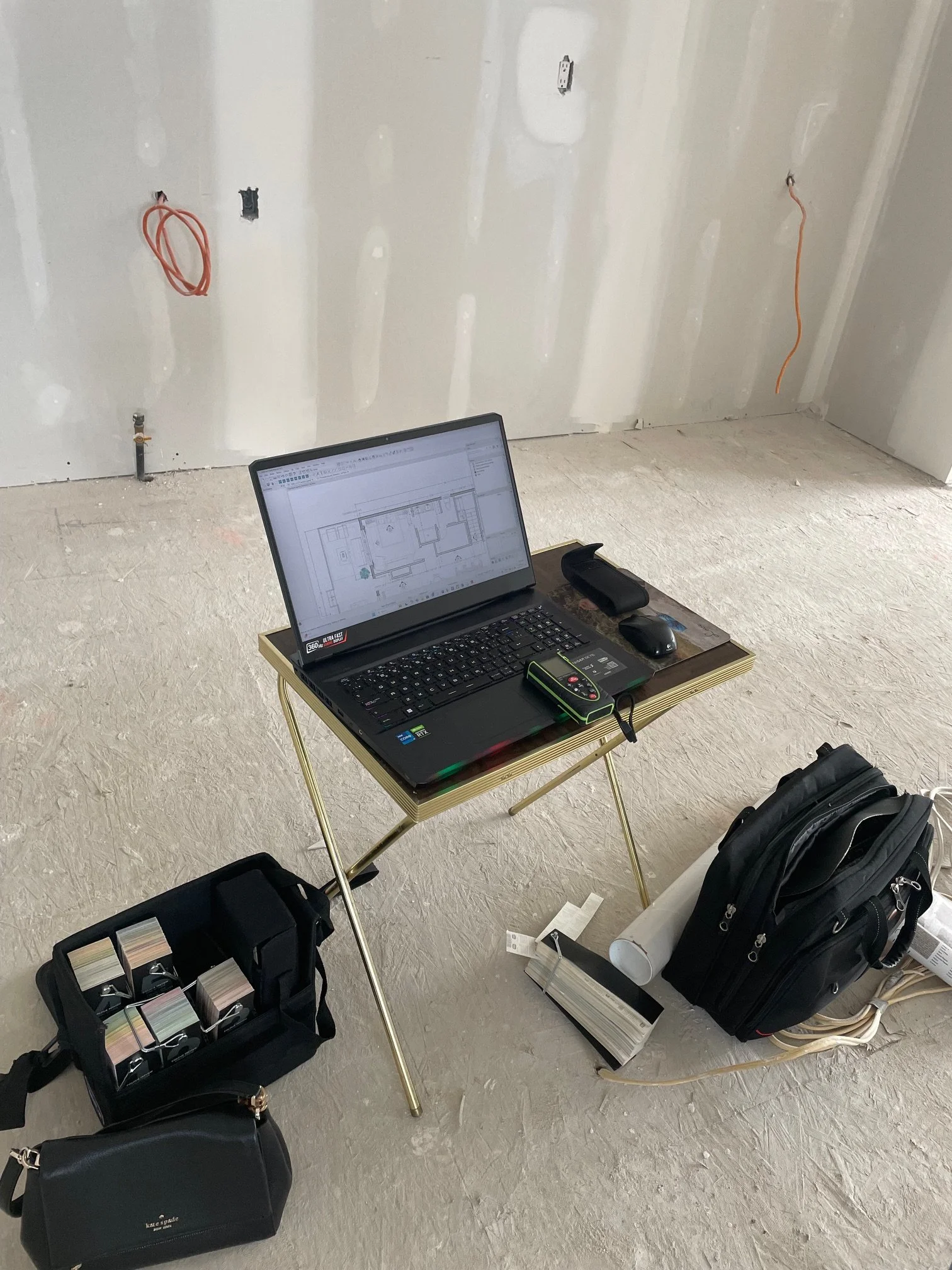 Laptop on a small table displaying a floor plan, surrounded by a backpack, a black bag, and office supplies, in a room under construction with unfinished walls and exposed electrical wiring.