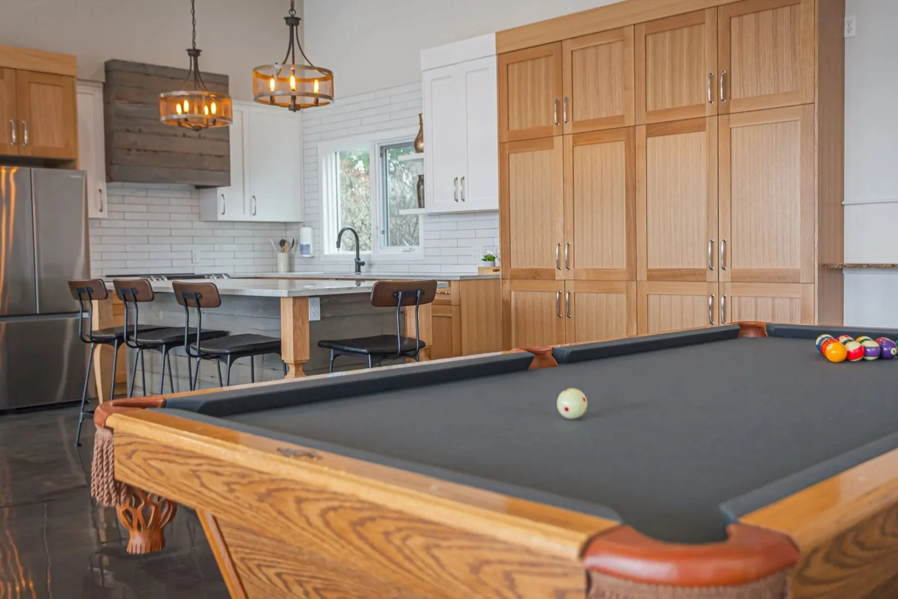 A modern kitchen with wooden cabinets, white brick backsplash, a sink window, and a kitchen island with bar stools. In the foreground, there is a pool table with a ball set and cue balls.
