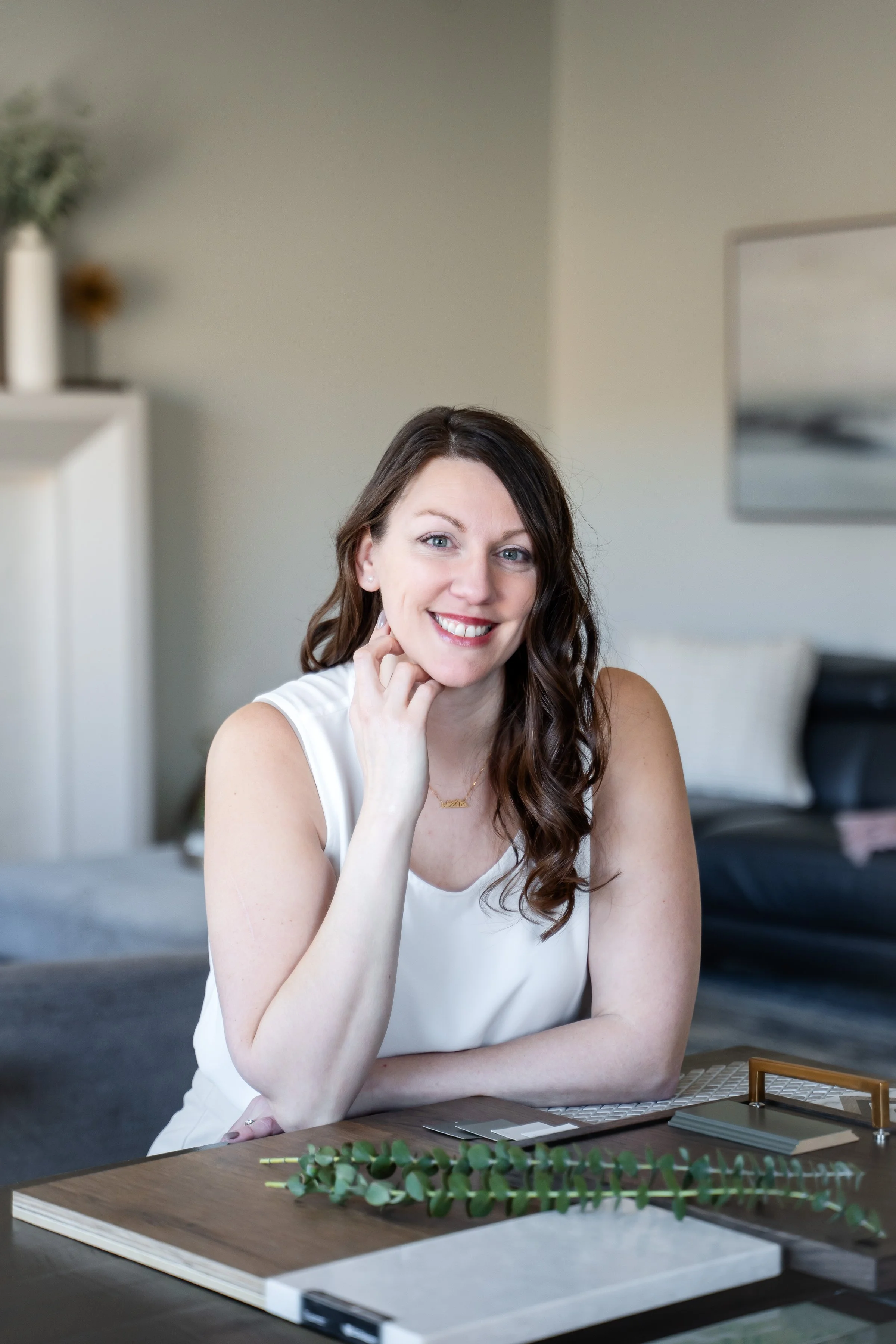 A woman with long, wavy brown hair, smiling, sitting at a table in a bright, modern living room, with plants and neutral-colored furniture in the background.