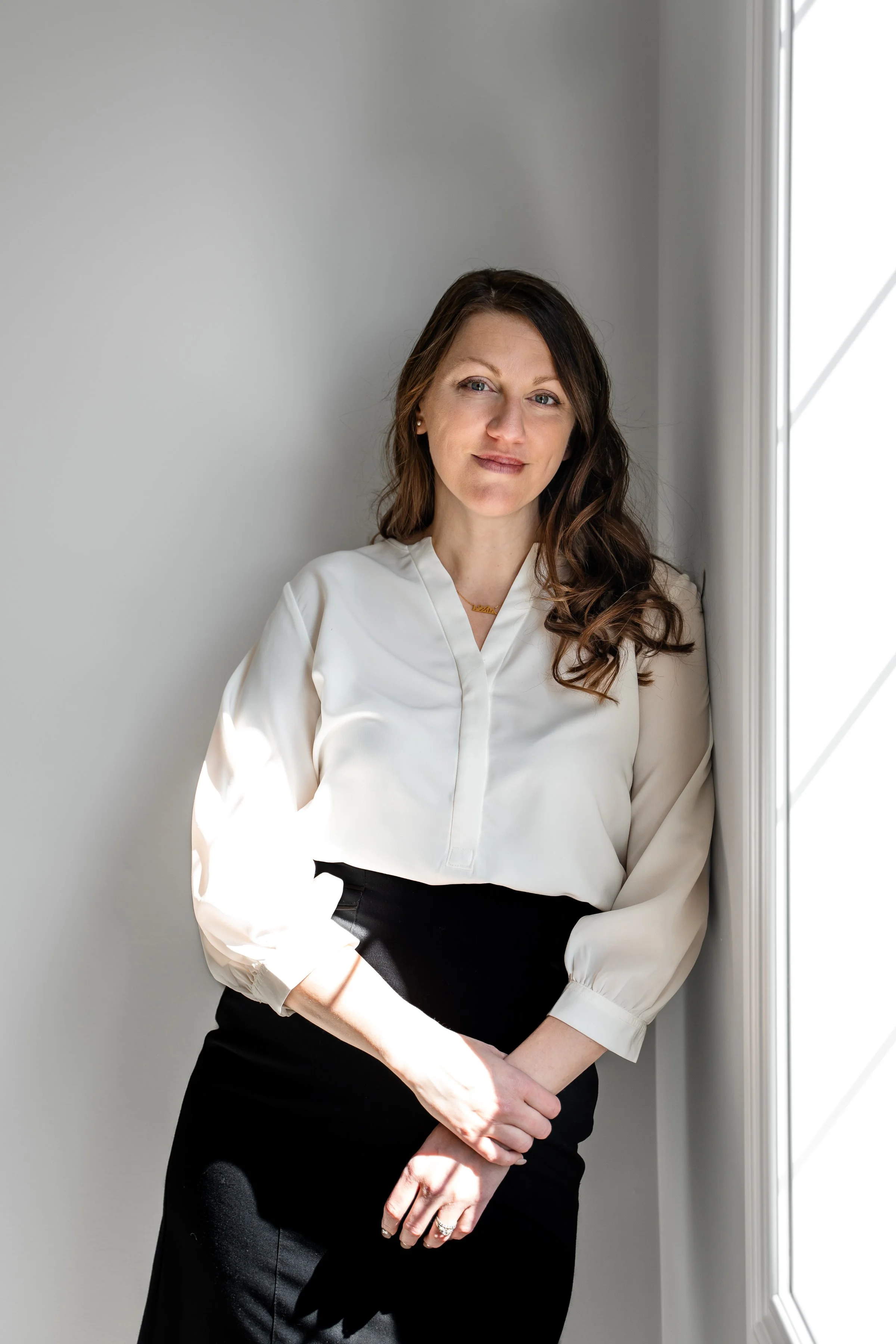 A woman leaning against a white wall near a window with natural light, wearing a white blouse and black skirt, with brown wavy hair, looking at the camera with a slight smile.