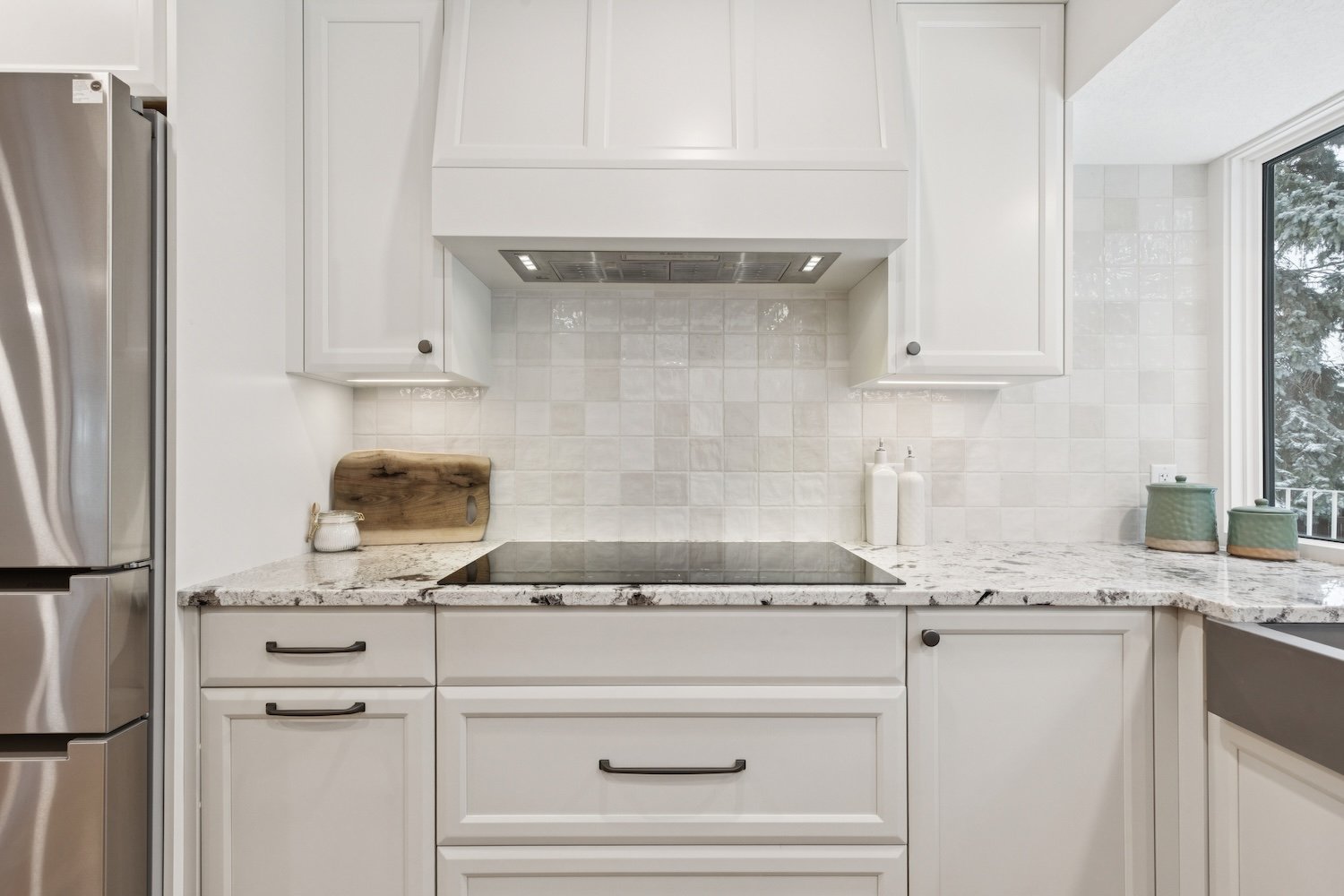 Modern white kitchen with granite countertop, stainless steel refrigerator, and decorative accessories near a window.