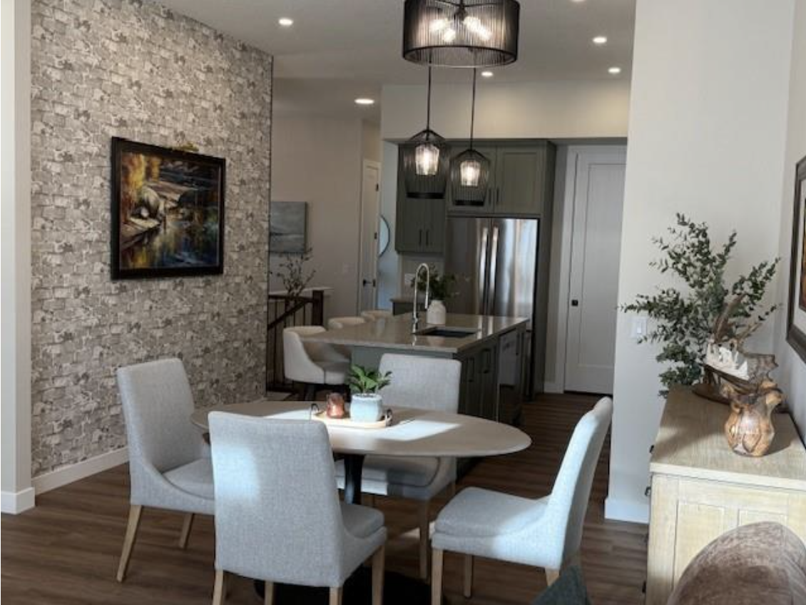 Living room and kitchen area with a stone accent wall, a round dining table with four light-colored chairs, herb plants, artwork, and a modern kitchen with gray cabinets and stainless steel appliances.