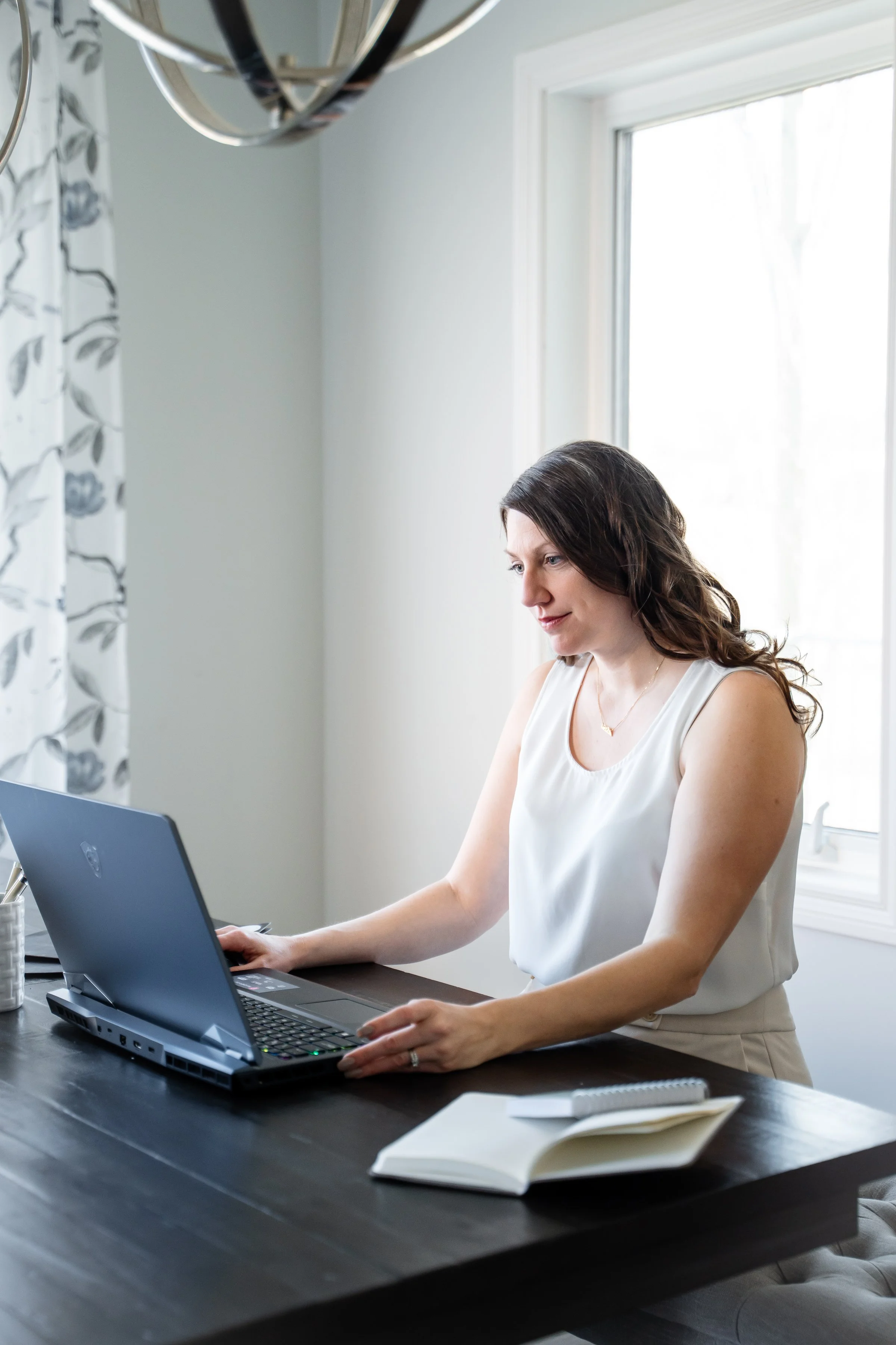 A woman with brown hair sitting at a black table working on a laptop, with an open notebook and a pen in front of her, beside a window with floral curtains.