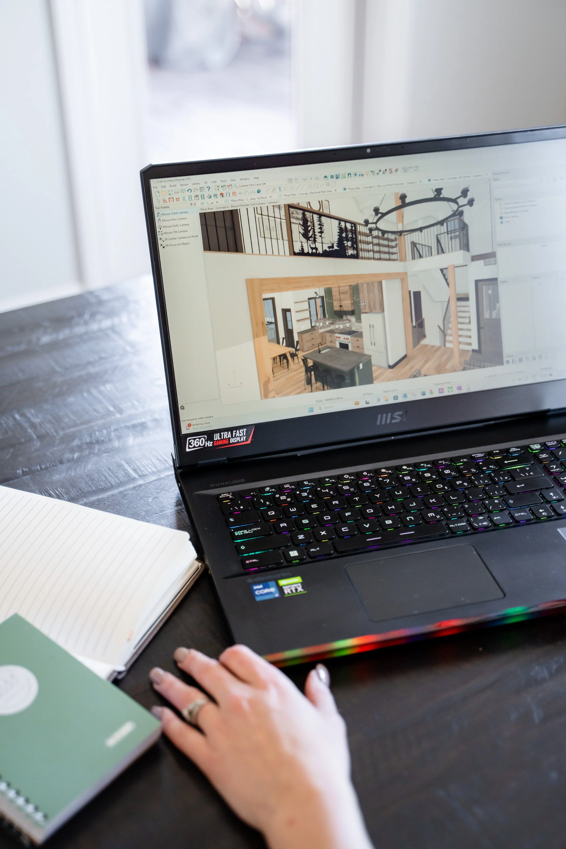 A person working on a laptop displaying a 3D home interior design, with a notebook nearby on a dark wooden table.