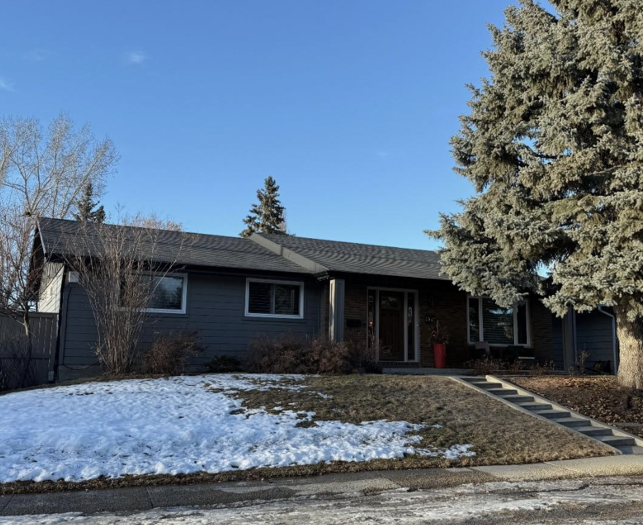 A single-story house with a gray exterior, black roof, and a small set of stairs leading to the front door. There is a large pine tree on the right, leafless bushes, patches of snow on the lawn, and a clear blue sky.