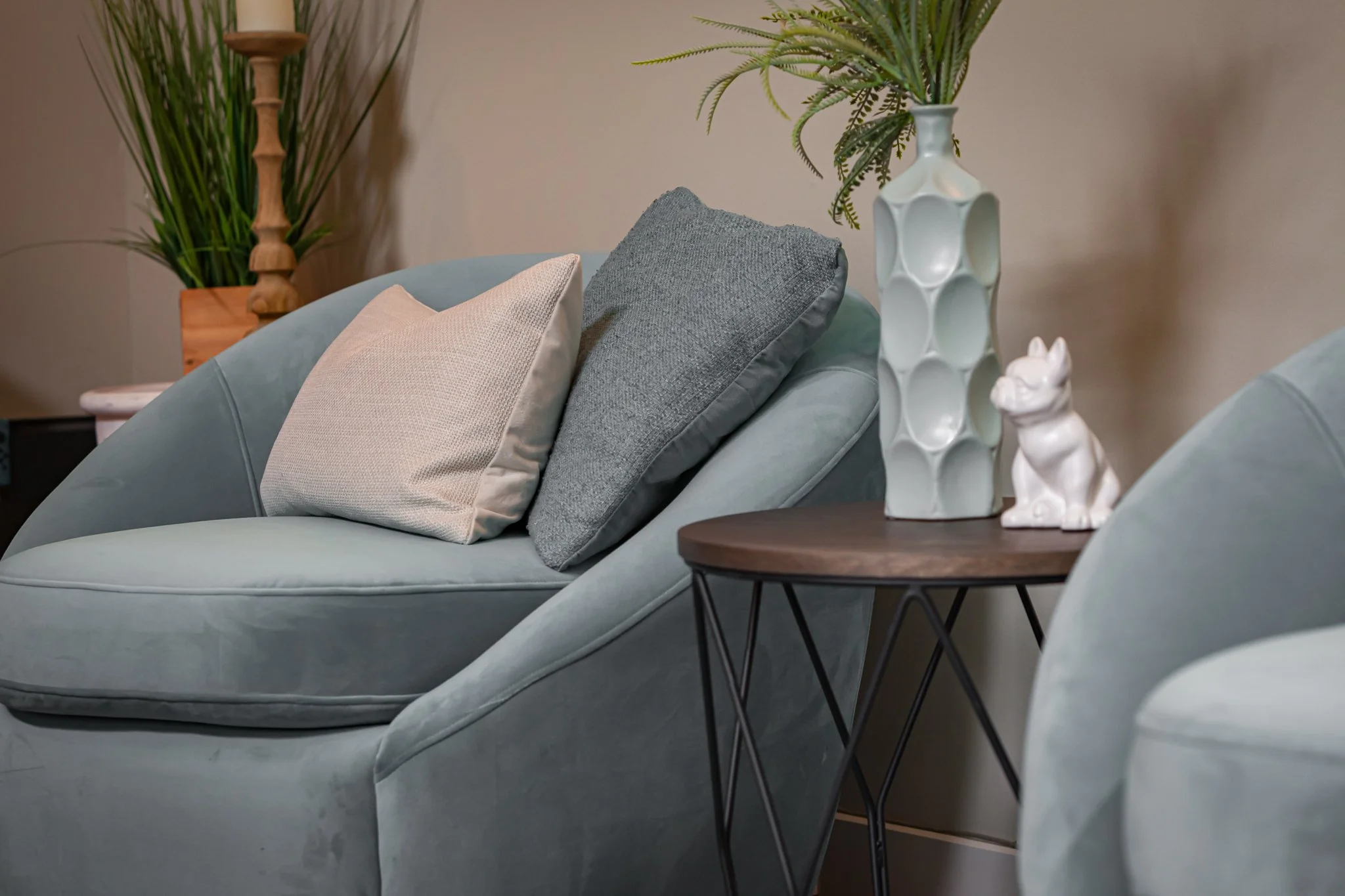 Living room with a light blue plush armchair, two throw pillows (one beige and one dark gray), a small round wooden side table with a decorative white ceramic vase and a small white ceramic dog figurine, and potted plants in the background.