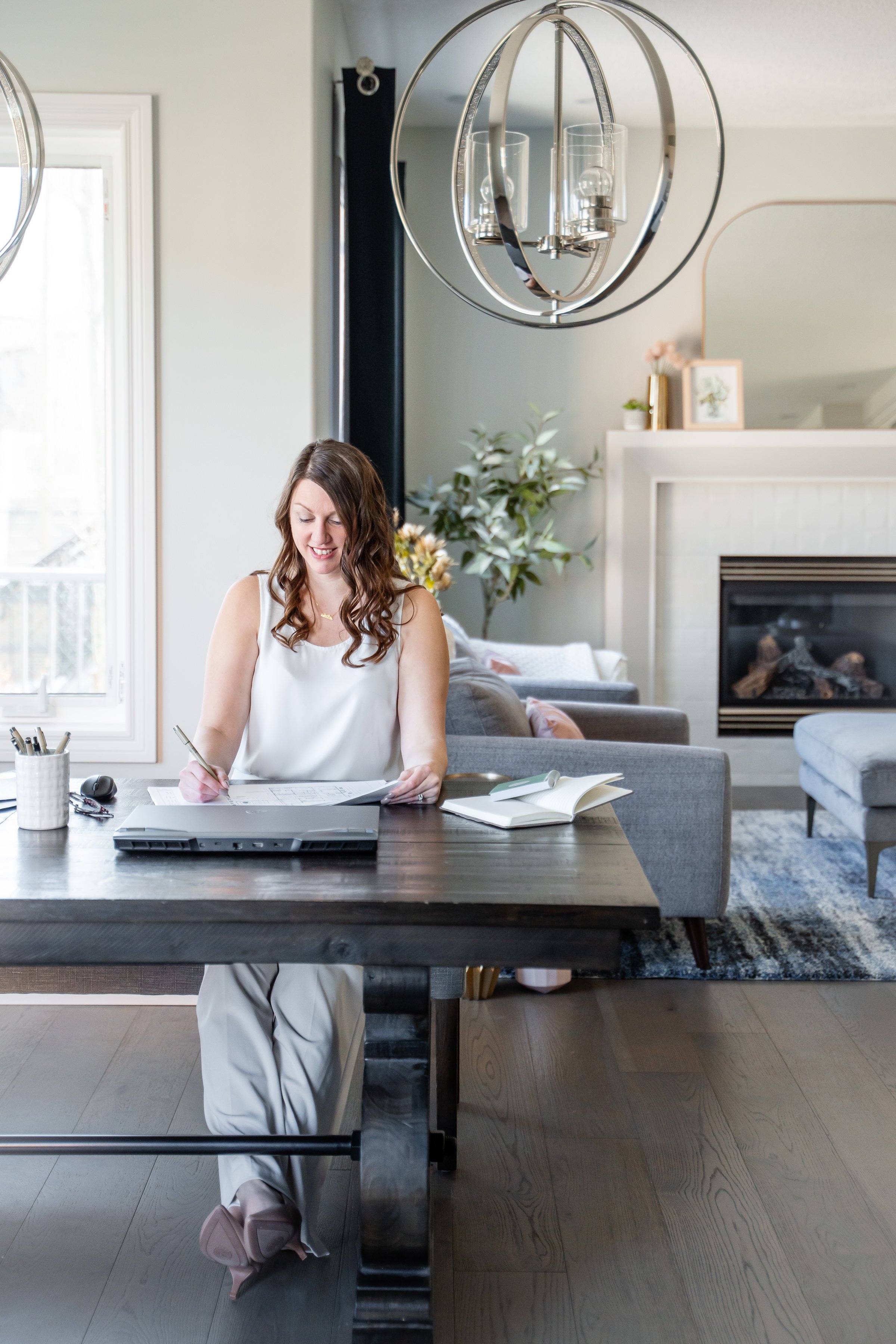 A woman sitting at a dark wooden table, working with a laptop and papers in a bright living room with a fireplace, gray sofa, and decorative greenery.