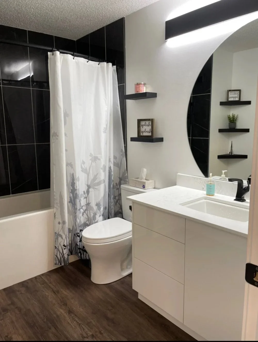 Photo of a modern bathroom with a black and white color scheme. Features include a shower with a white floral shower curtain, a white toilet, a white vanity with a sink, a large round mirror, and three black wall shelves holding decor items. The floor is wood-like laminate.