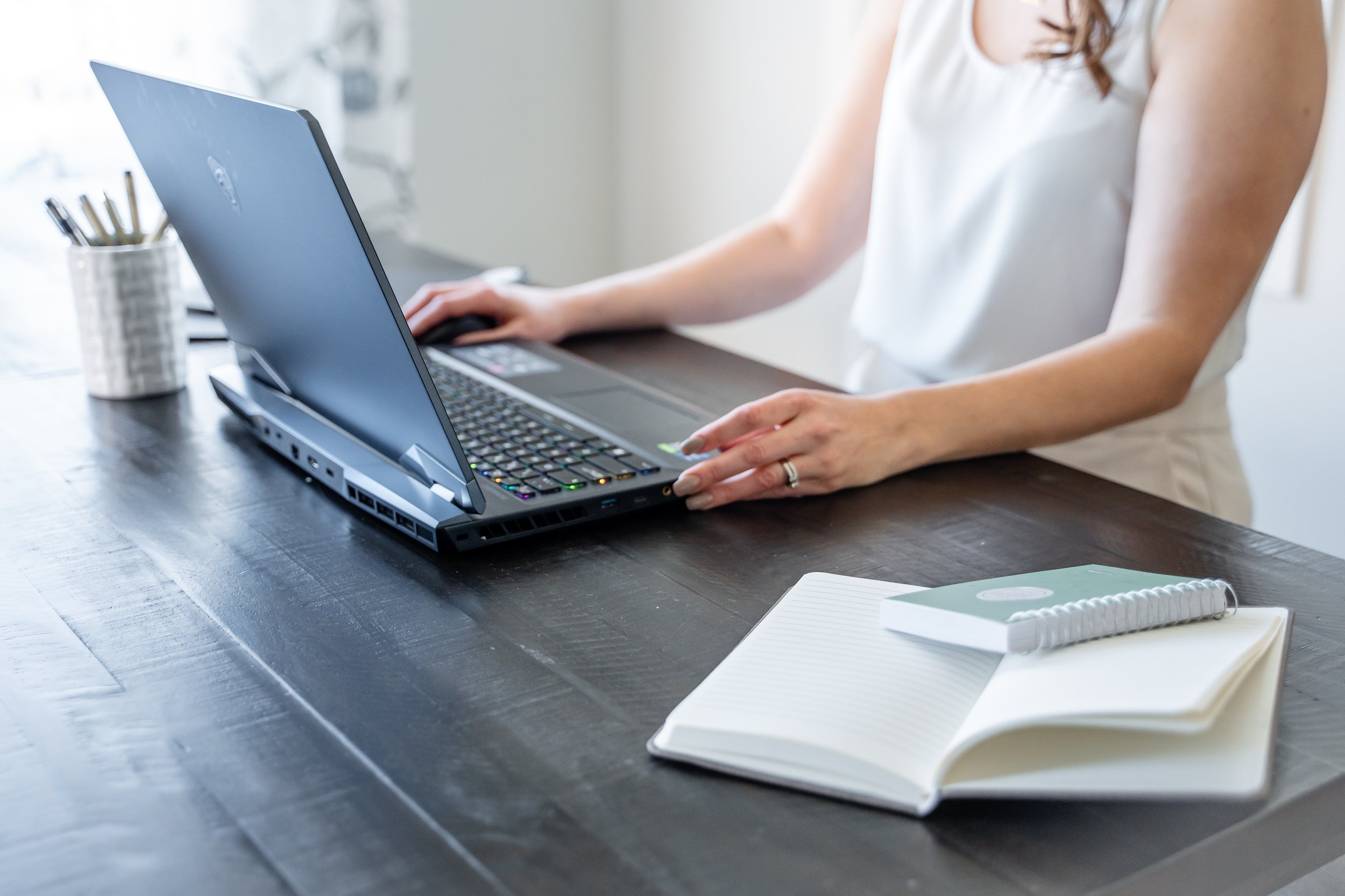 A woman in a white sleeveless top working on a laptop at a dark wooden desk, with a notebook and phone nearby.