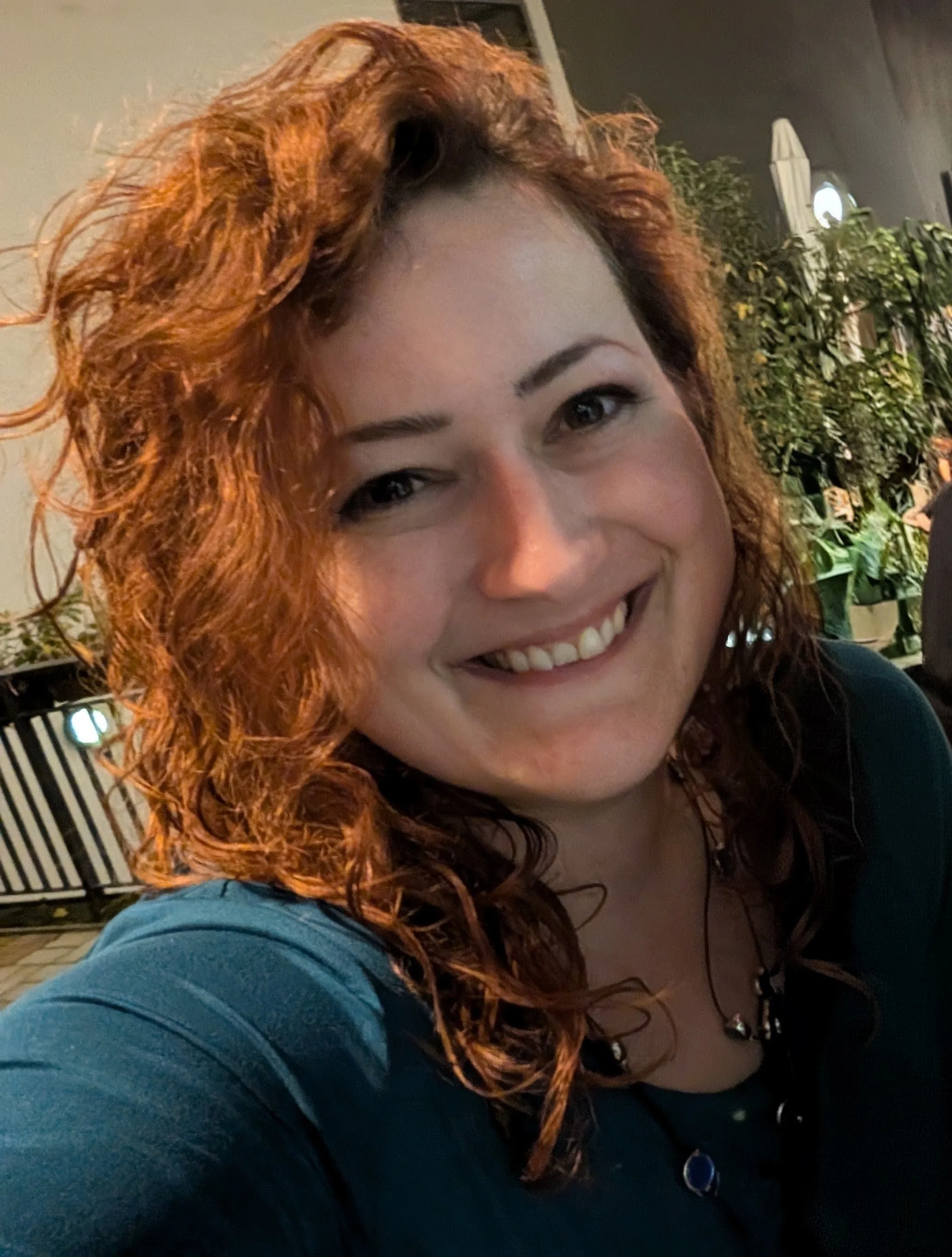 Ein Porträt einer lächelnden Frau mit lockigem, rotem Haar, die in einem Restaurant oder einer Bar sitzt, im Hintergrund sind Pflanzen und Lichter sichtbar.