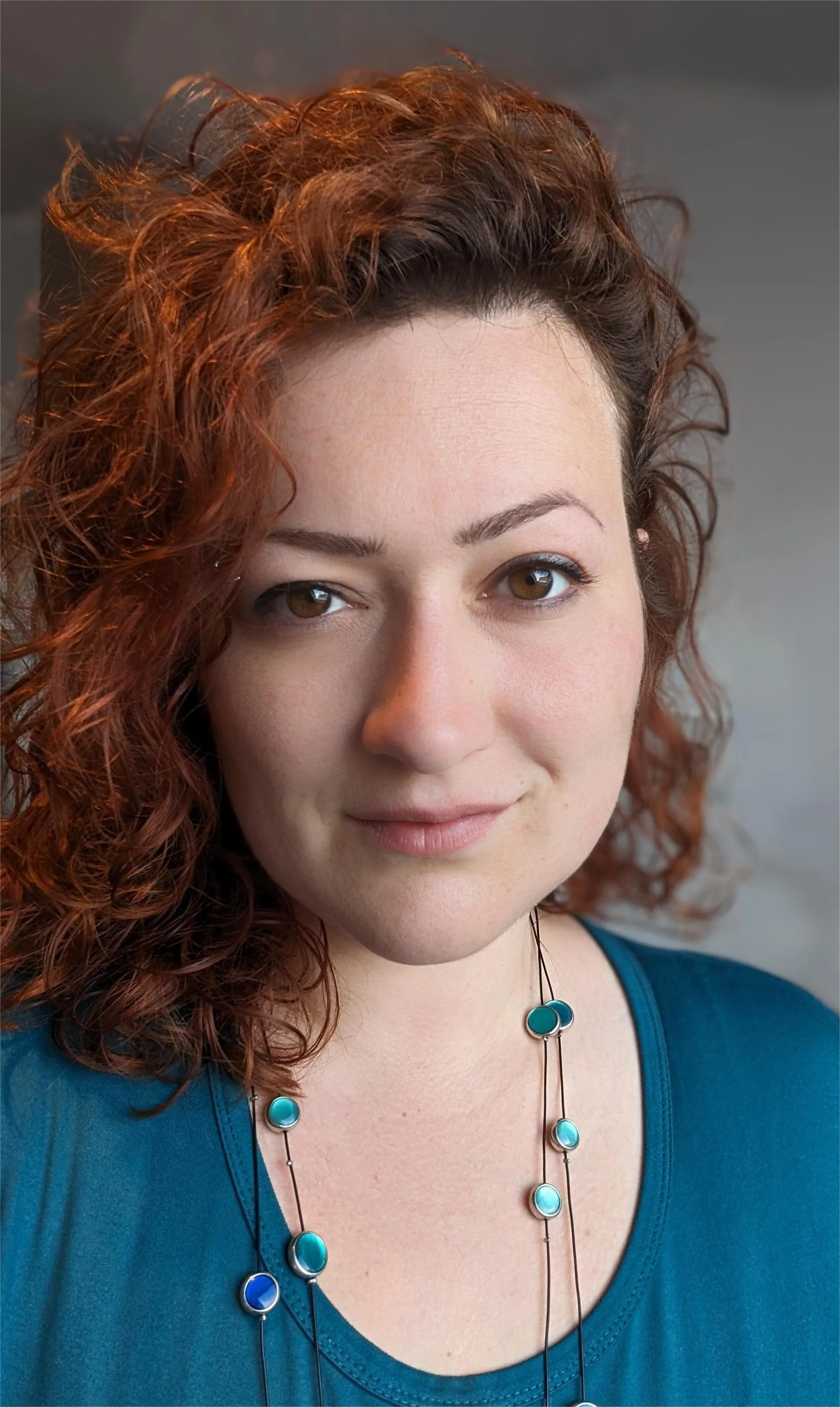 Nahaufnahme eines Lächelns einer jungen Frau mit lockigem rotbraunem Haar, braunen Augen und heller Haut, trägt eine blaue Bluse und eine lange Halskette mit blauen und türkisen Anhängern.