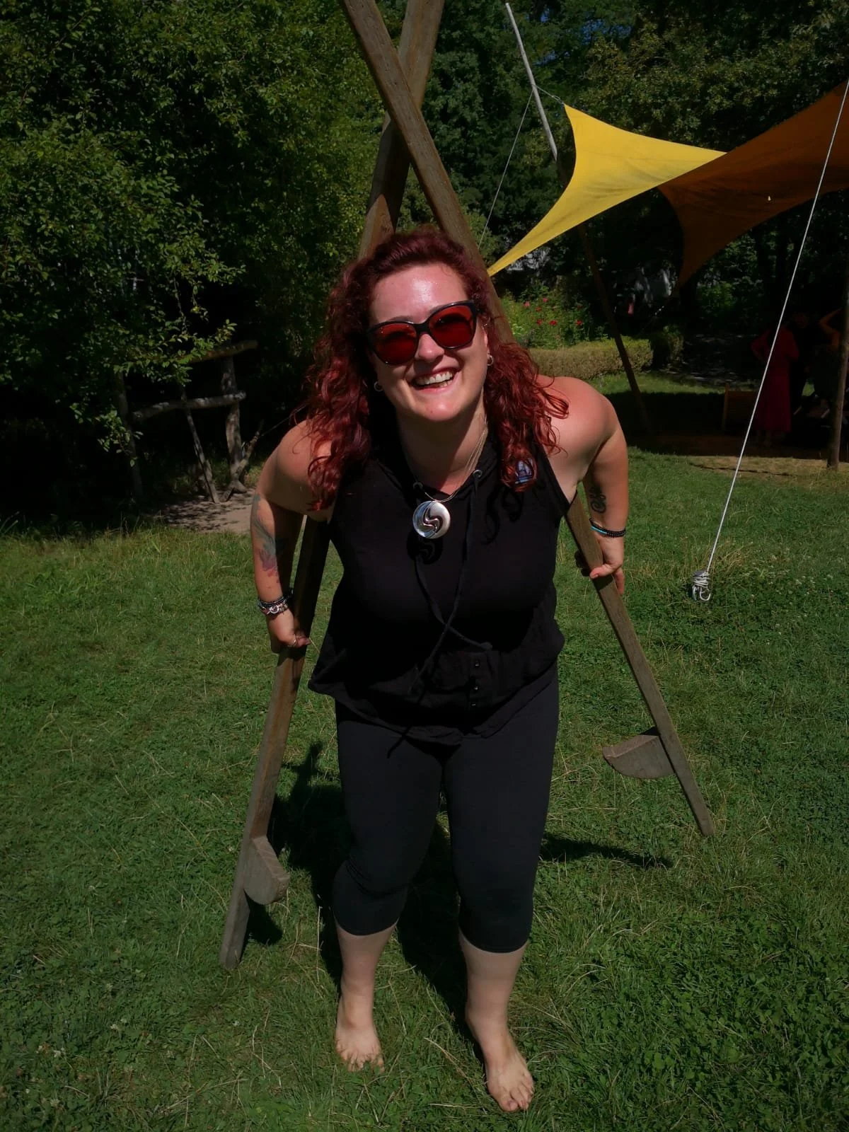 Eine lachende Frau mit roten Haaren trägt Sonnenbrille, schwarzes Top, schwarze Kapuzenhose, steht barfuß auf Gras, hält ein Holzgestell im Rücken, im Hintergrund bunte Sonnensegel und Bäume.