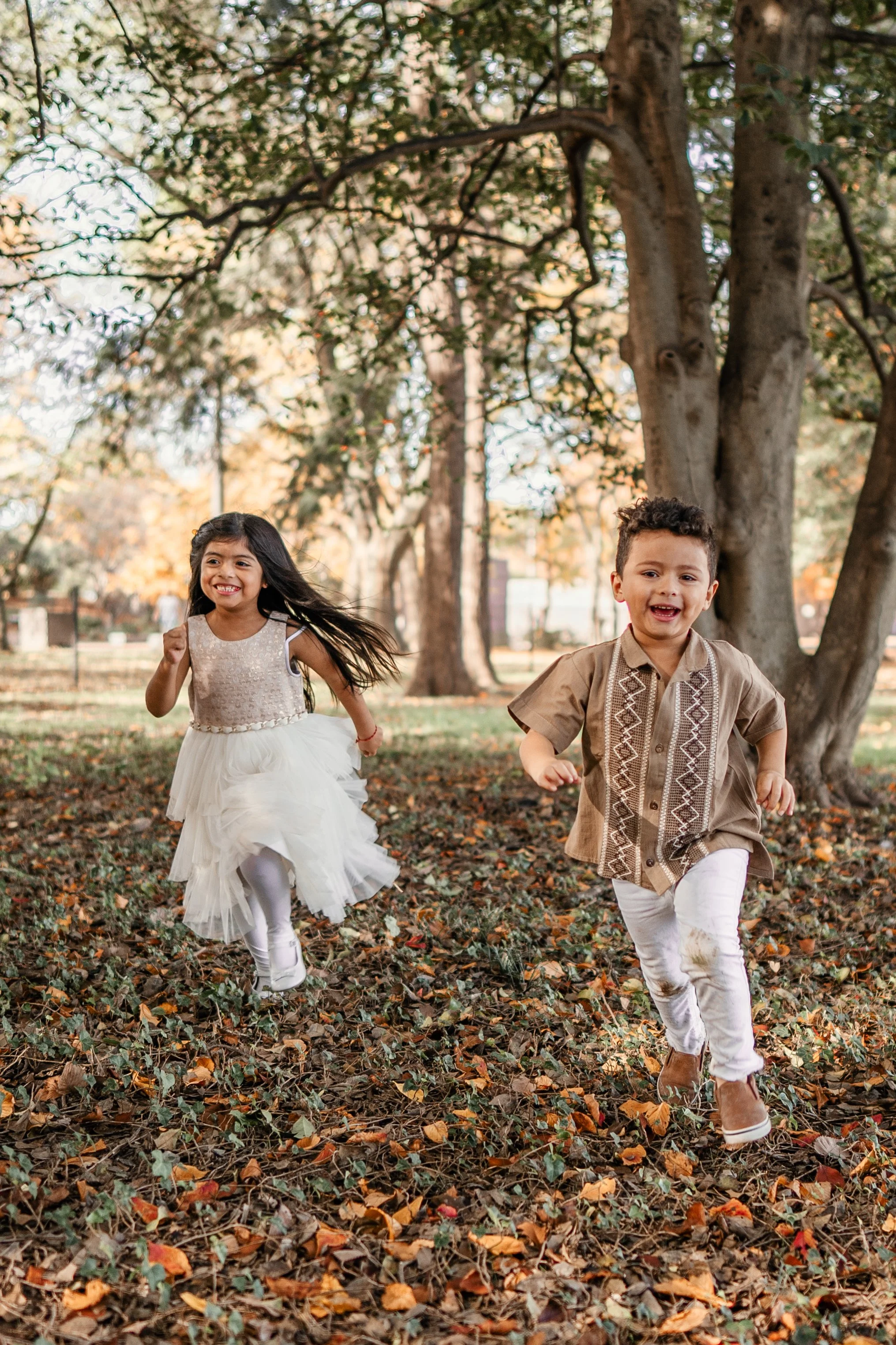 Two young children, a girl in a white dress and a boy in a tan shirt, running and playing outdoors in a park with autumn leaves on the ground and trees in the background.