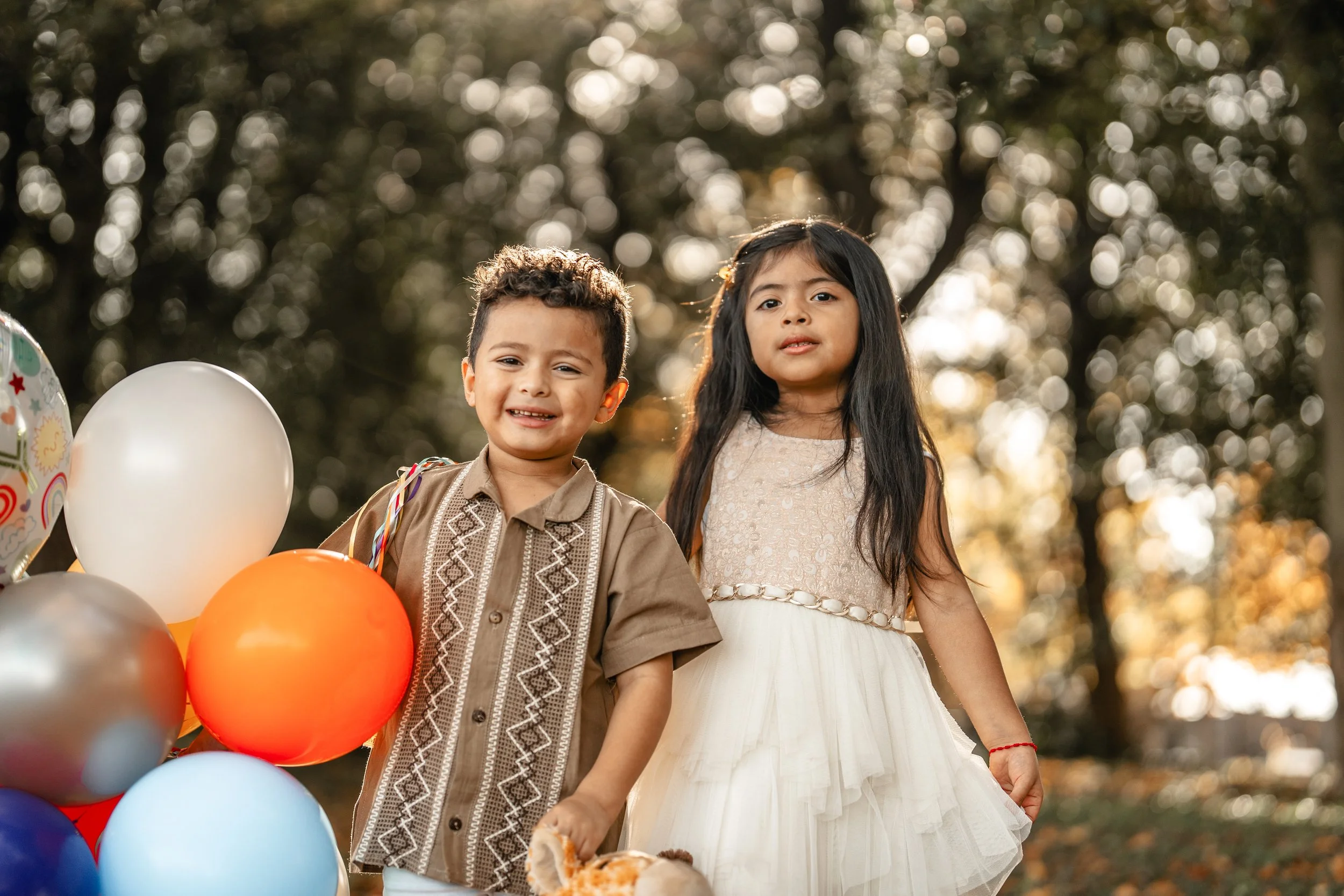 Two children, a boy and a girl, standing outdoors surrounded by balloons, with trees in the background and warm sunlight.