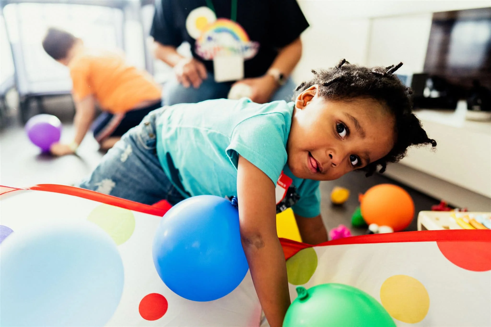 A young boy crawling on the floor with colorful balloons in front of him, with other children and an adult in the background during a playroom activity.