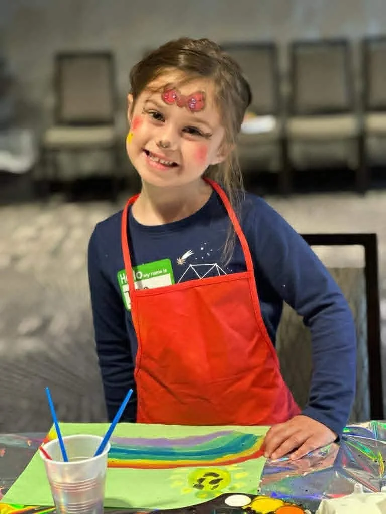 A young girl smiling, with face paint and a colorful apron, standing next to a table with a rainbow-colored painting and art supplies, in an indoor setting.