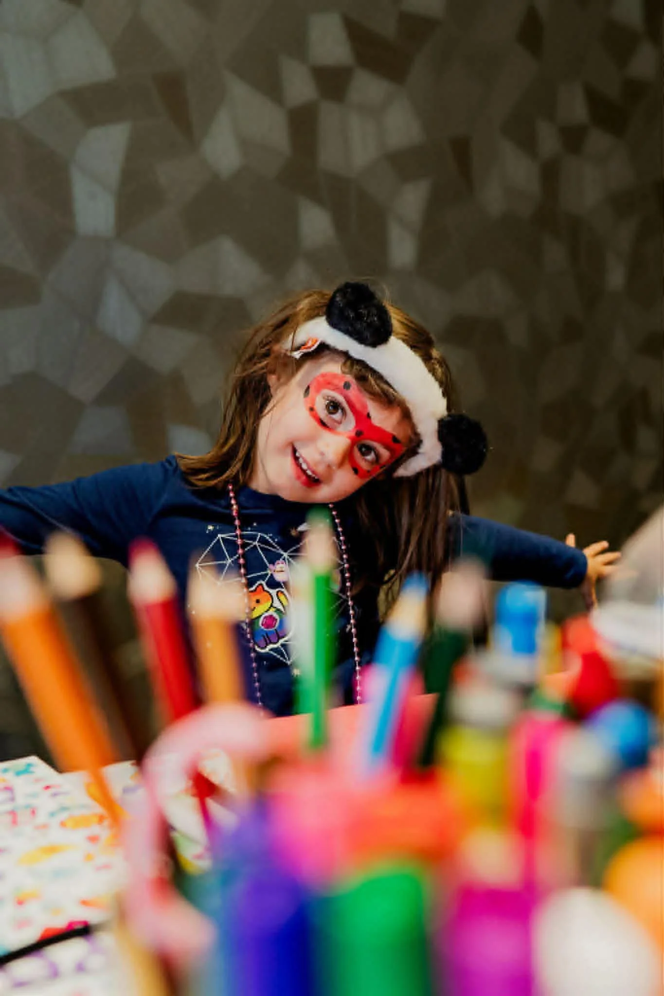 A young girl wearing a panda hat, red glasses, and colorful beaded necklace, smiling with arms outstretched, surrounded by colorful markers and art supplies