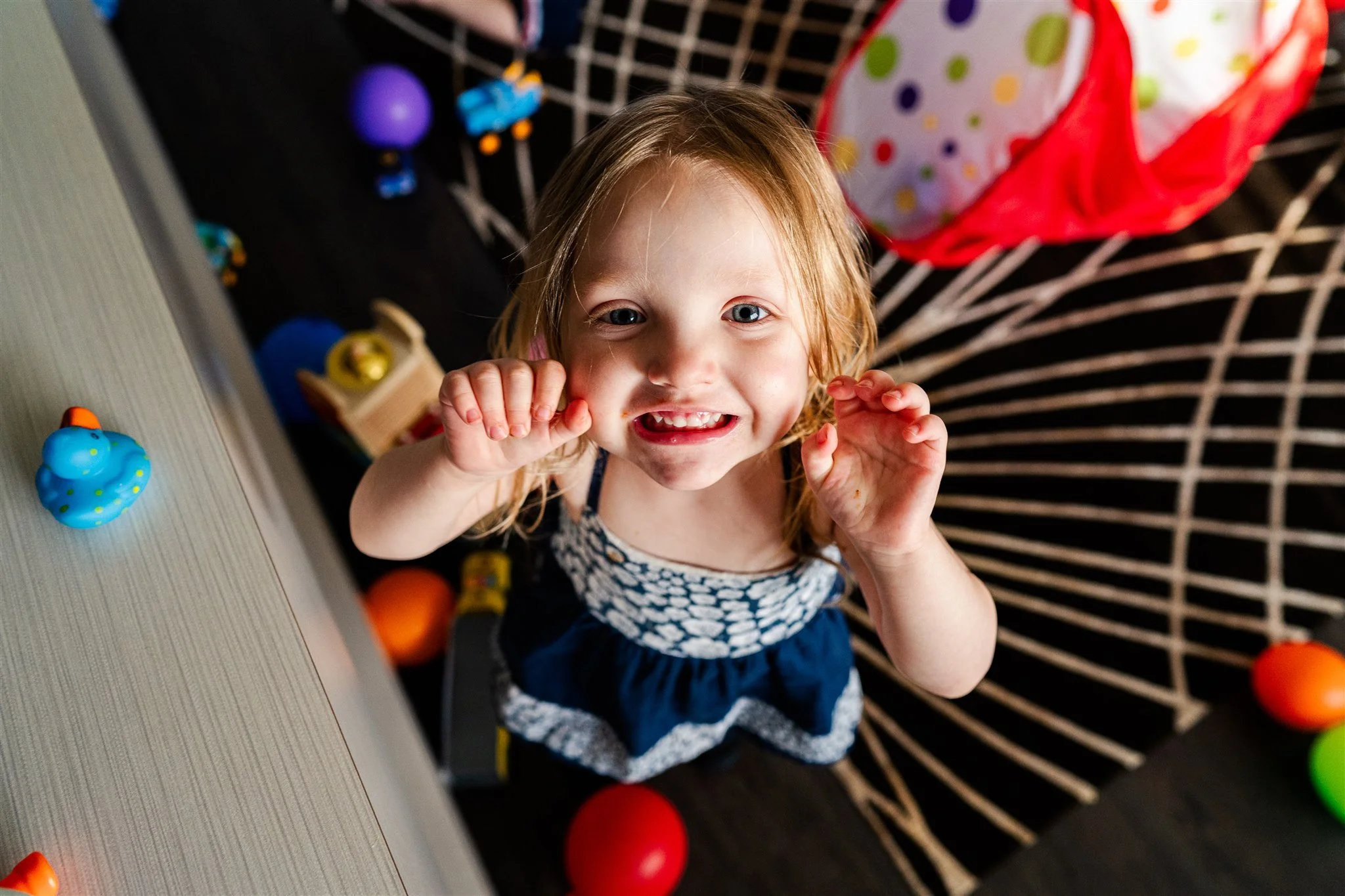 A young girl with blonde hair smiling at the camera, raising her hands in a playful gesture, surrounded by colorful toys and balls on the floor.