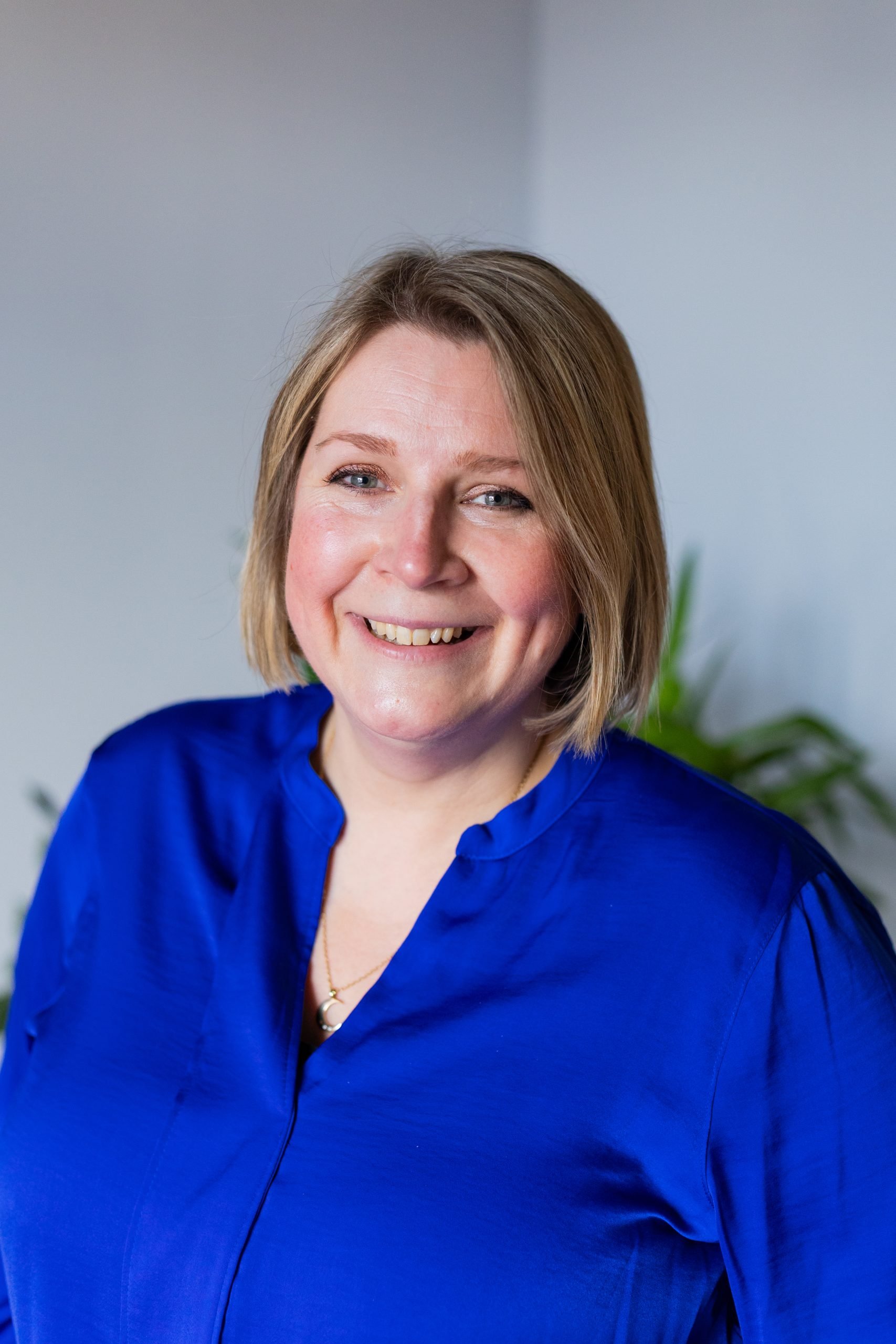 Close-up of Jacqui of Neon & Co smiling with short blonde hair, wearing a royal blue blouse, in front of a light grey background with a blurred green plant.