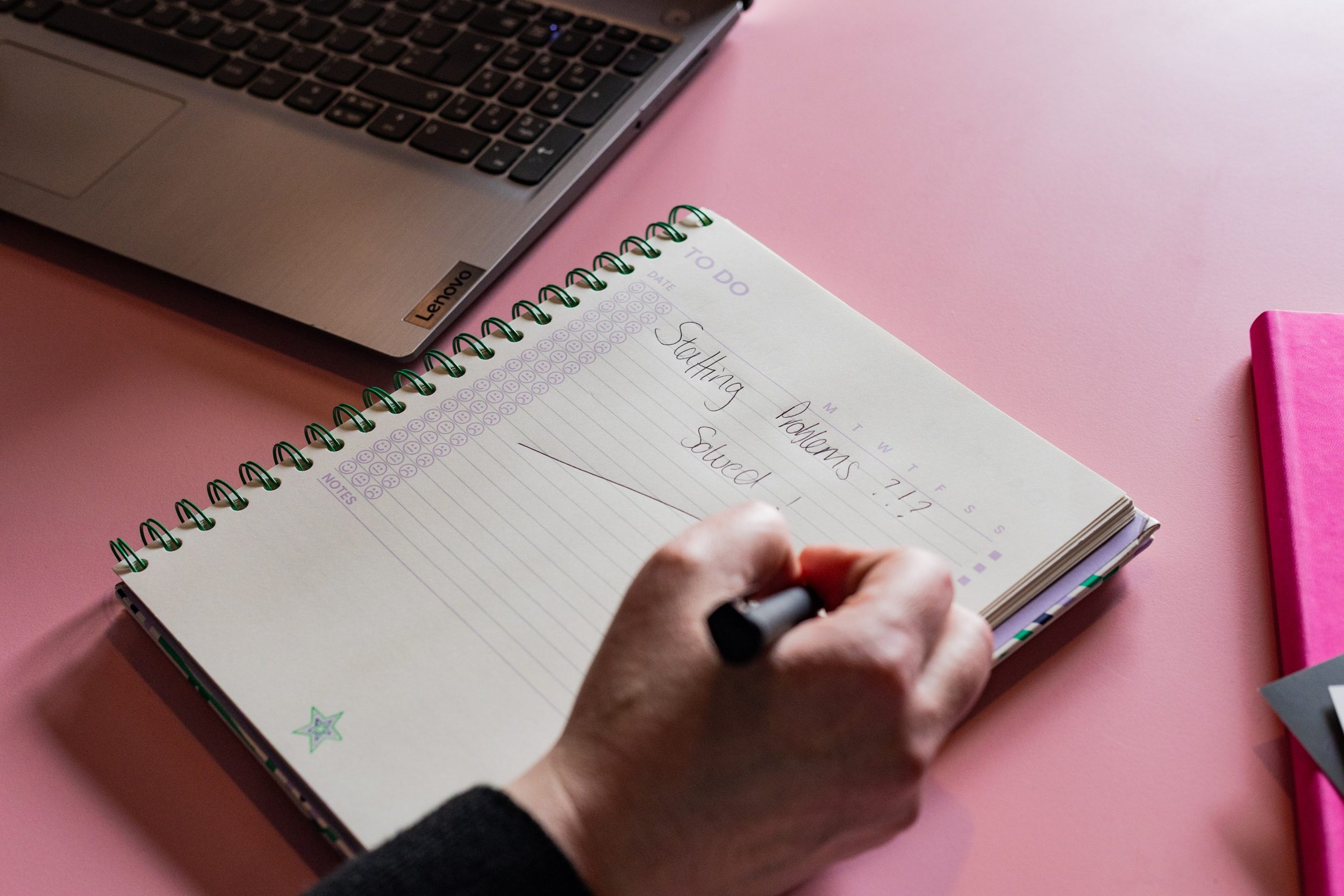 A person writing in a spiral notebook titled 'To Do' with handwritten text 'Starting Problems Solved.' The notebook has electronic smiley faces and a star decoration. A laptop is partially visible on a pink surface.