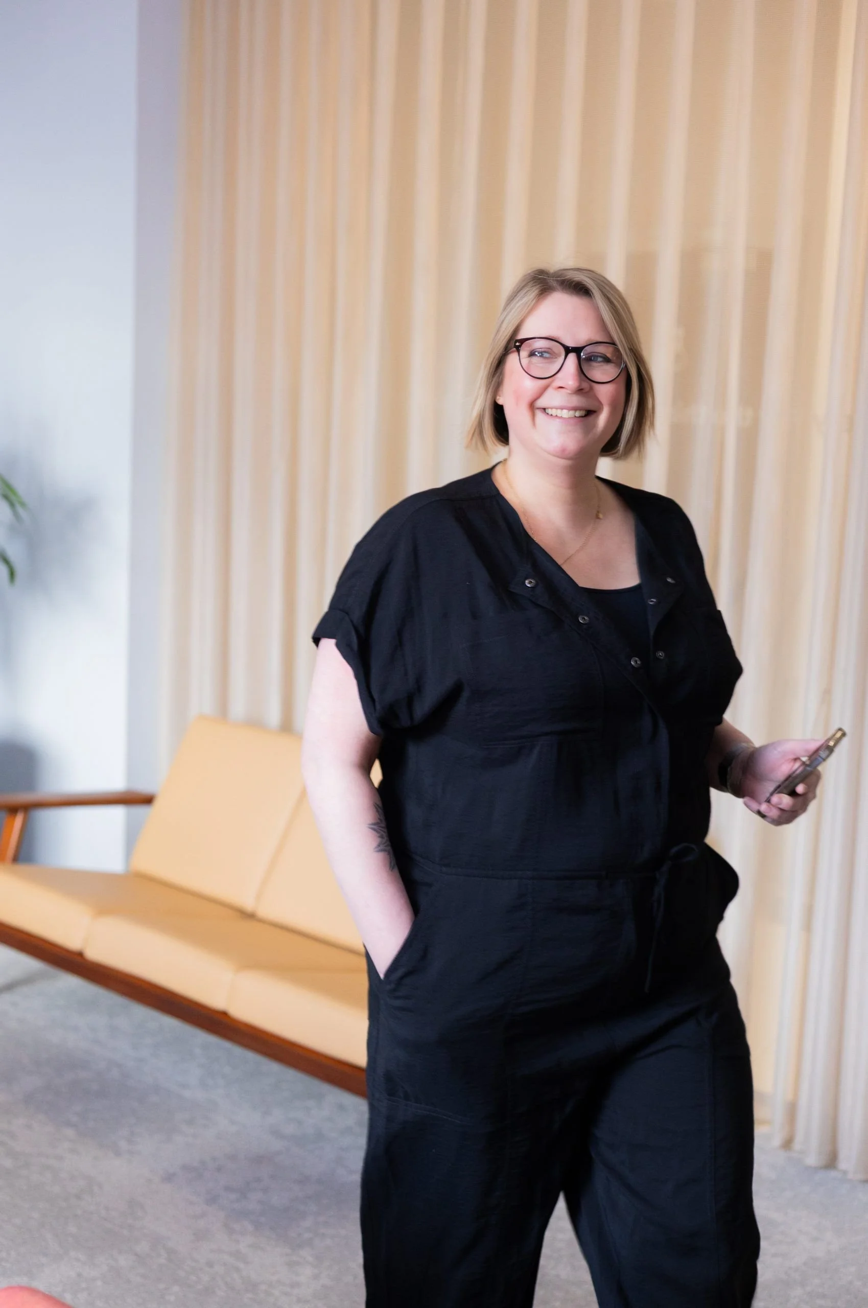 A woman with glasses and short blond hair smiling, standing indoors in front of beige curtains and a mustard-colored bench, holding a phone