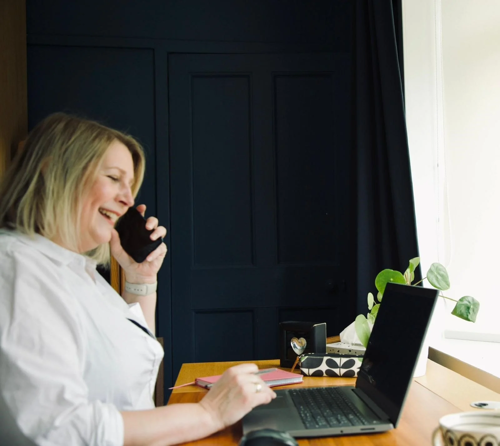 Jacqui of Neon & Co, with blonde hair, wearing a white shirt, is sitting at a desk, smiling and talking on a smartphone. She is working on a laptop, with a pink notebook and a plant on the desk.