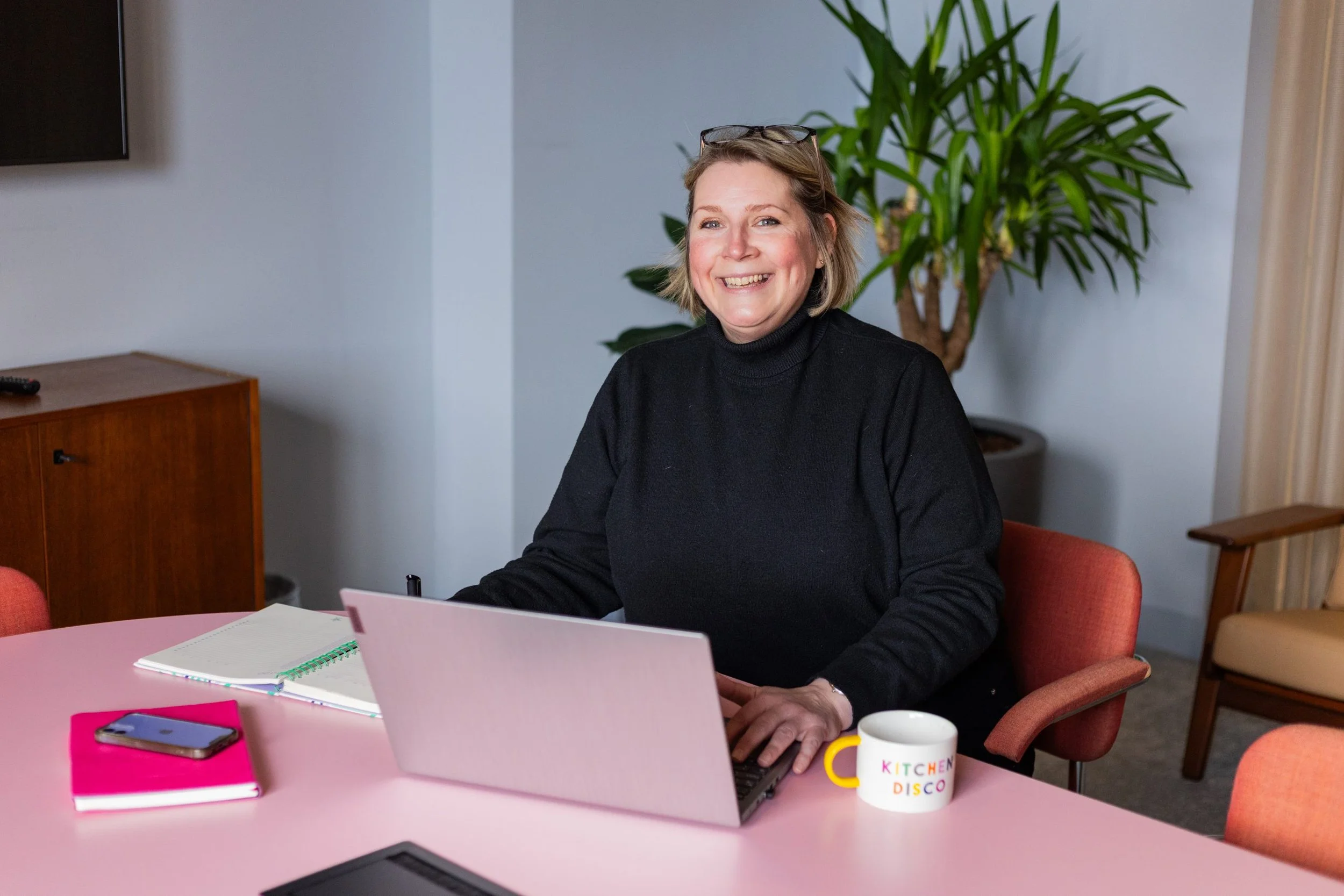 A smiling woman with short blond hair wearing glasses on her head, sitting at a pink table with a laptop, notebooks, a phone, and a mug that says 'KITCHEN DISCO' in colorful letters. There's a large green potted plant in the background.