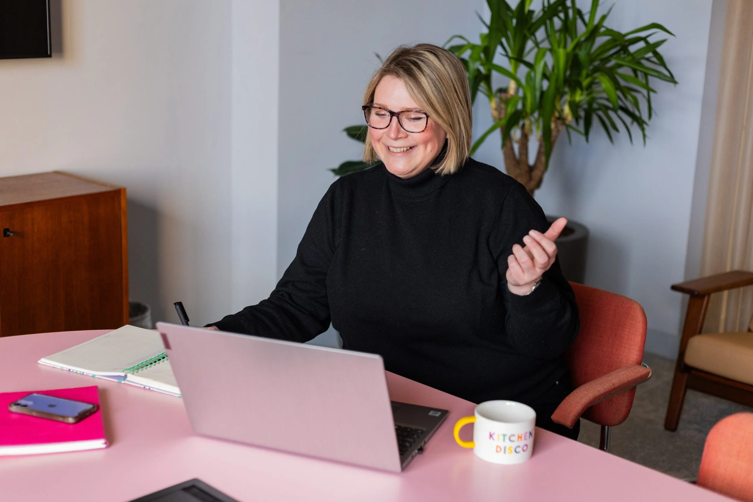 A woman with glasses and blonde hair smiling while sitting at a pink table with notebooks, a laptop, a smartphone, and a coffee mug that says 'Kitchen Disco.' She appears to be in a meeting or working in a modern office with a large green plant in the background.