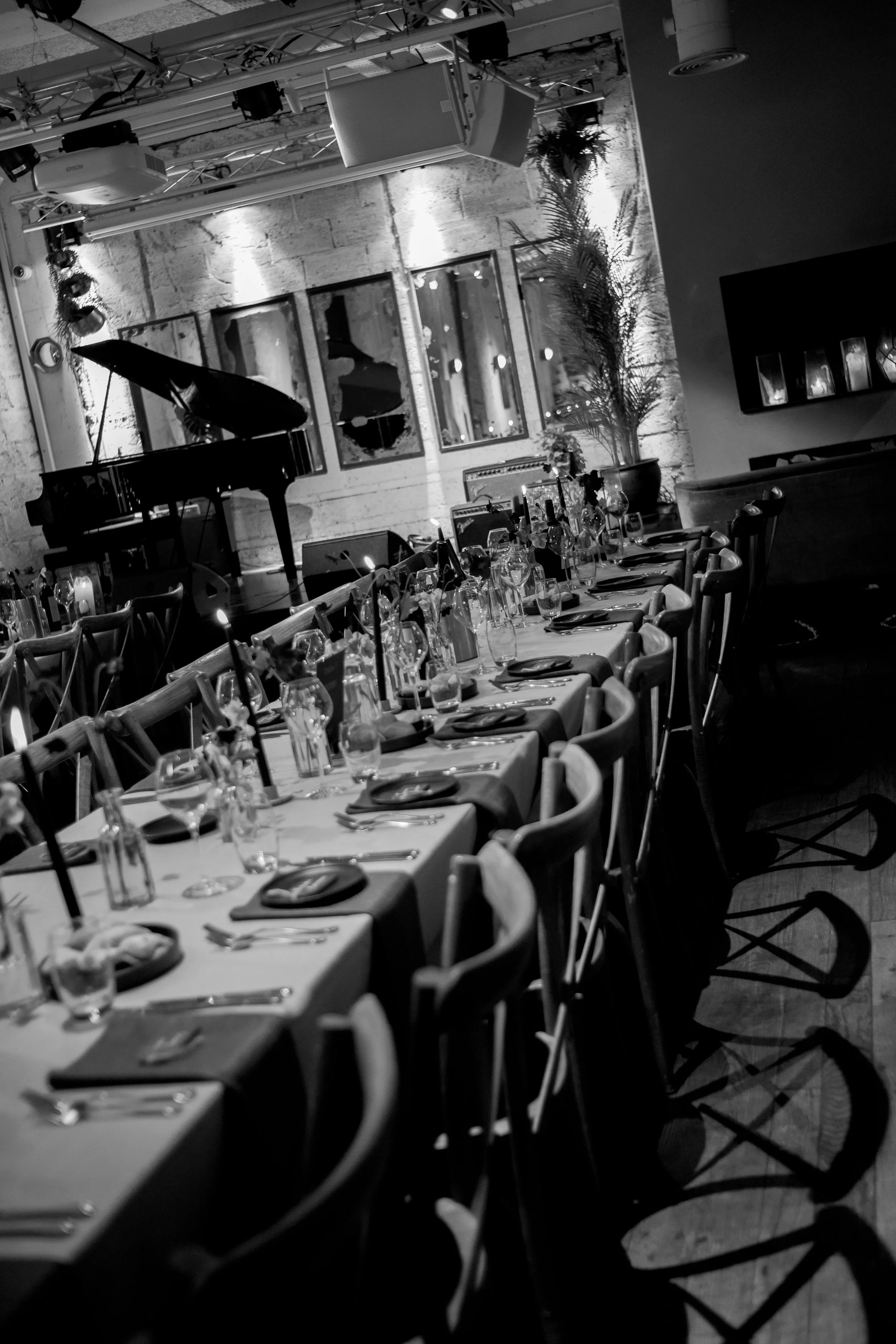 A dinner event setup with a long table, chairs, glasses, and plates in an indoor venue with a piano, artwork, and potted plant.